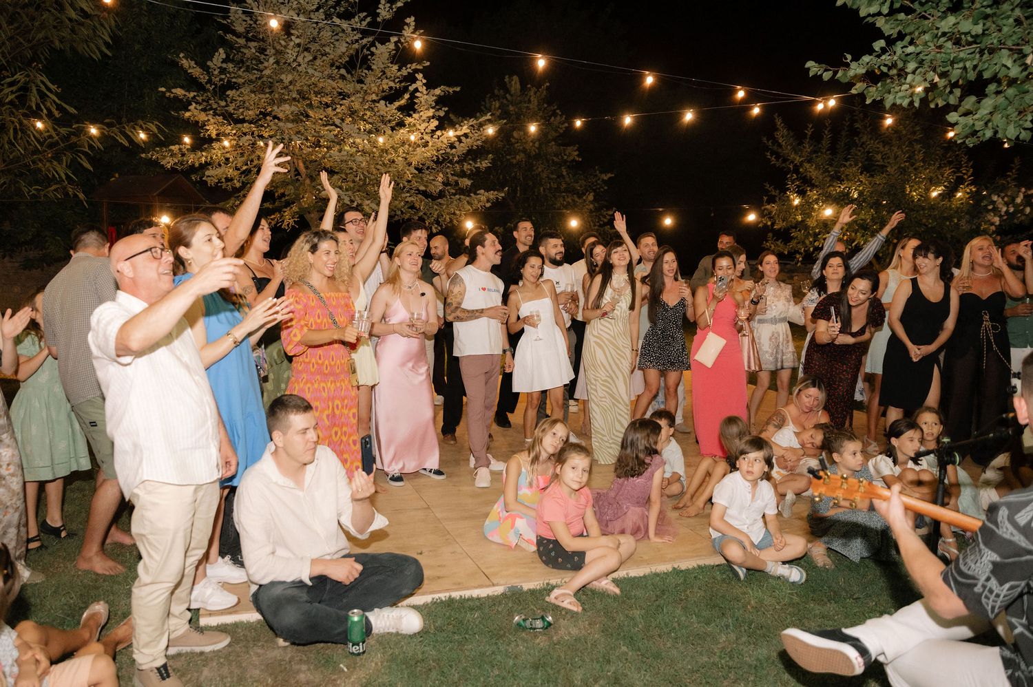 People dance and celebrate at an outdoor evening party under string lights and trees.