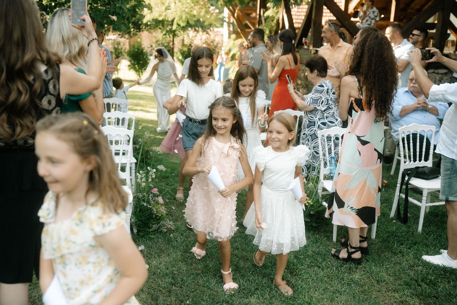 Children in white dresses dance and play together at an outdoor garden party celebration.