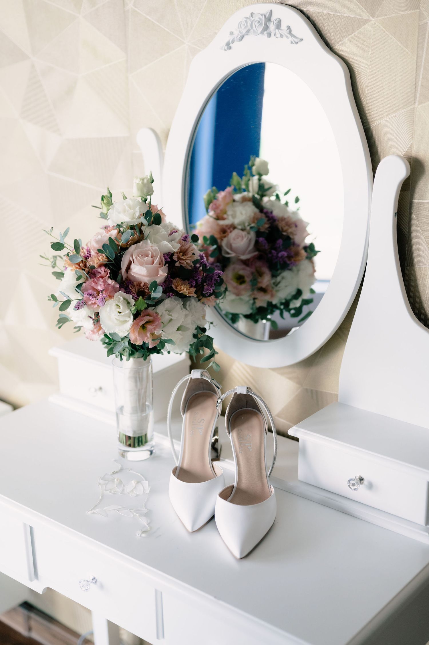 White wedding shoes and delicate floral bouquet arranged with round mirrors on a white vanity table.
