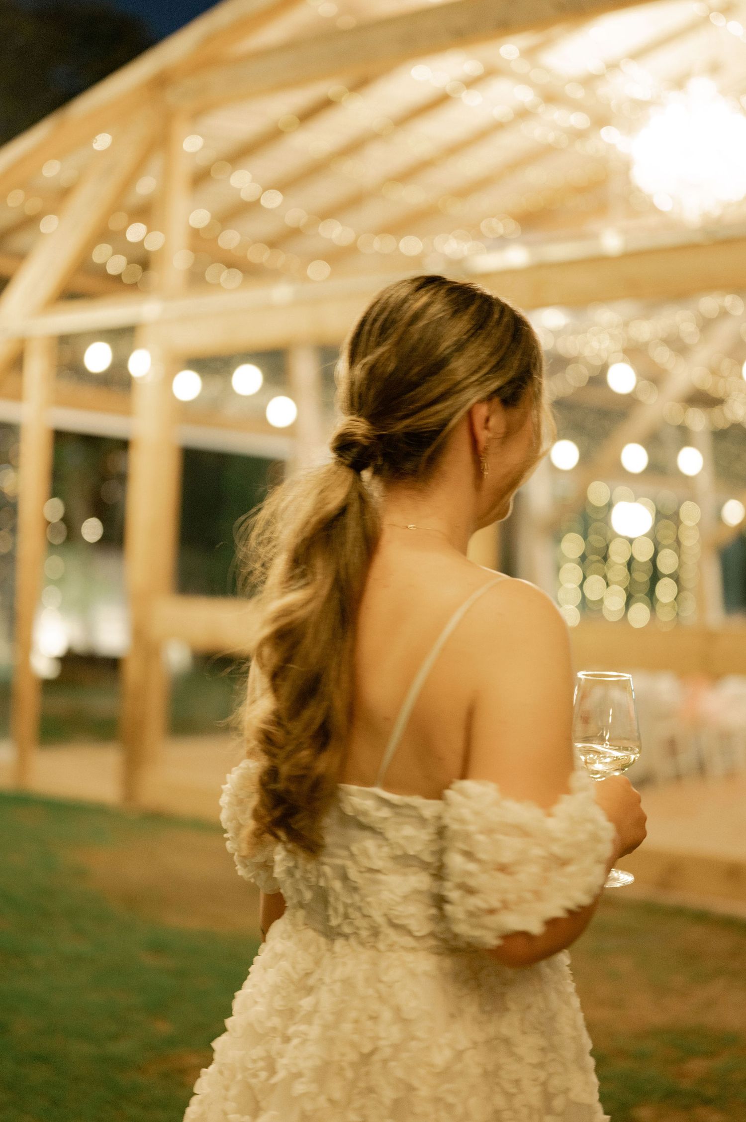 A bride in a white wedding dress with ruffled details dances under twinkling lights at an outdoor evening reception.