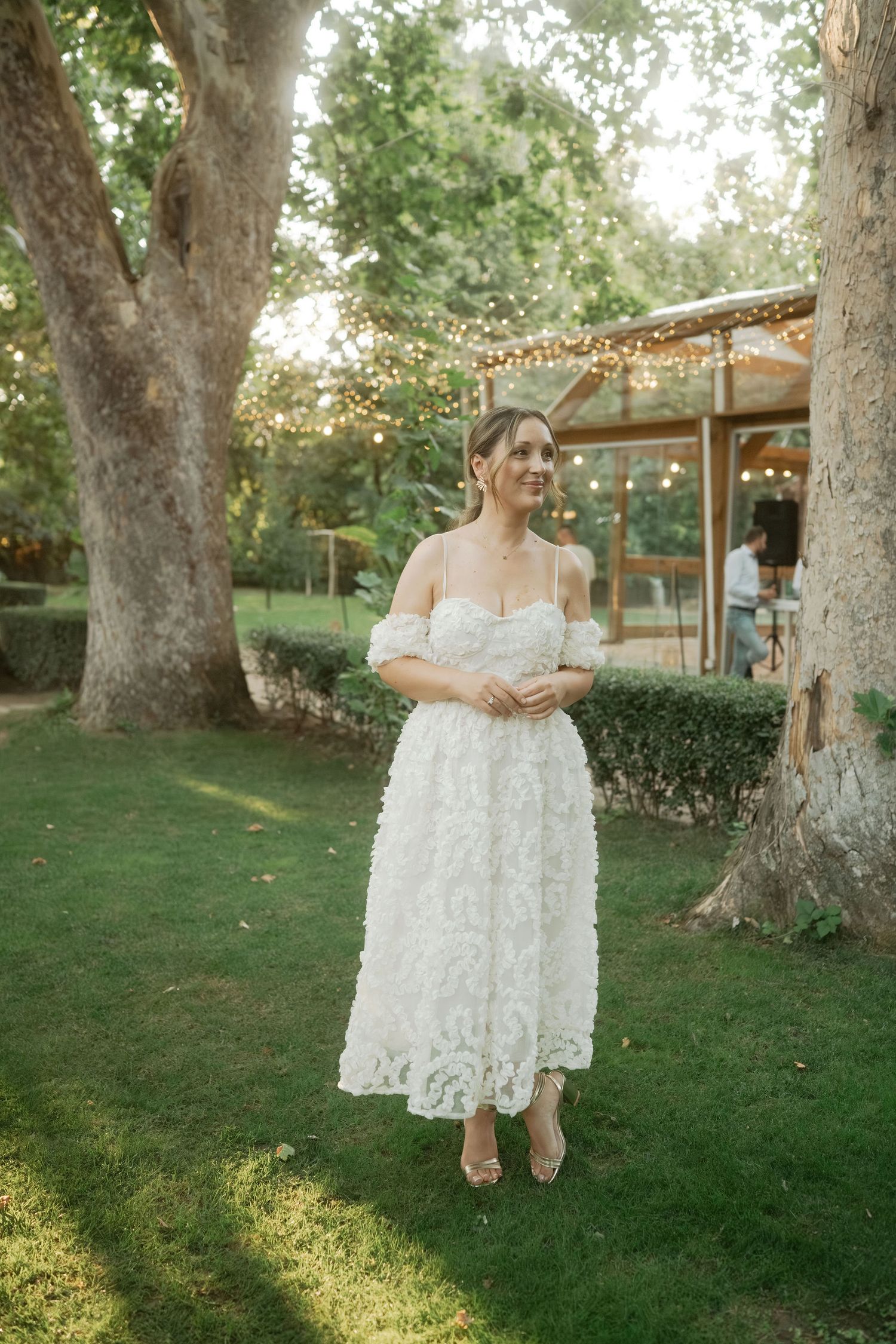 A white lace dress with intricate detailing is showcased in a garden setting with string lights and mature trees.