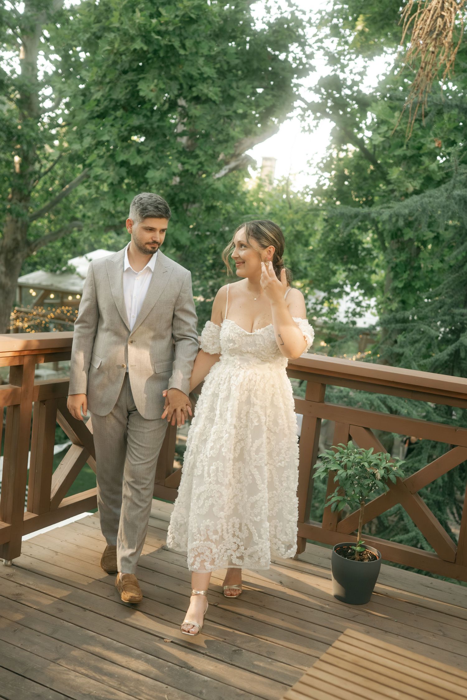 A couple walks hand in hand on a wooden deck wearing elegant wedding attire surrounded by lush greenery.