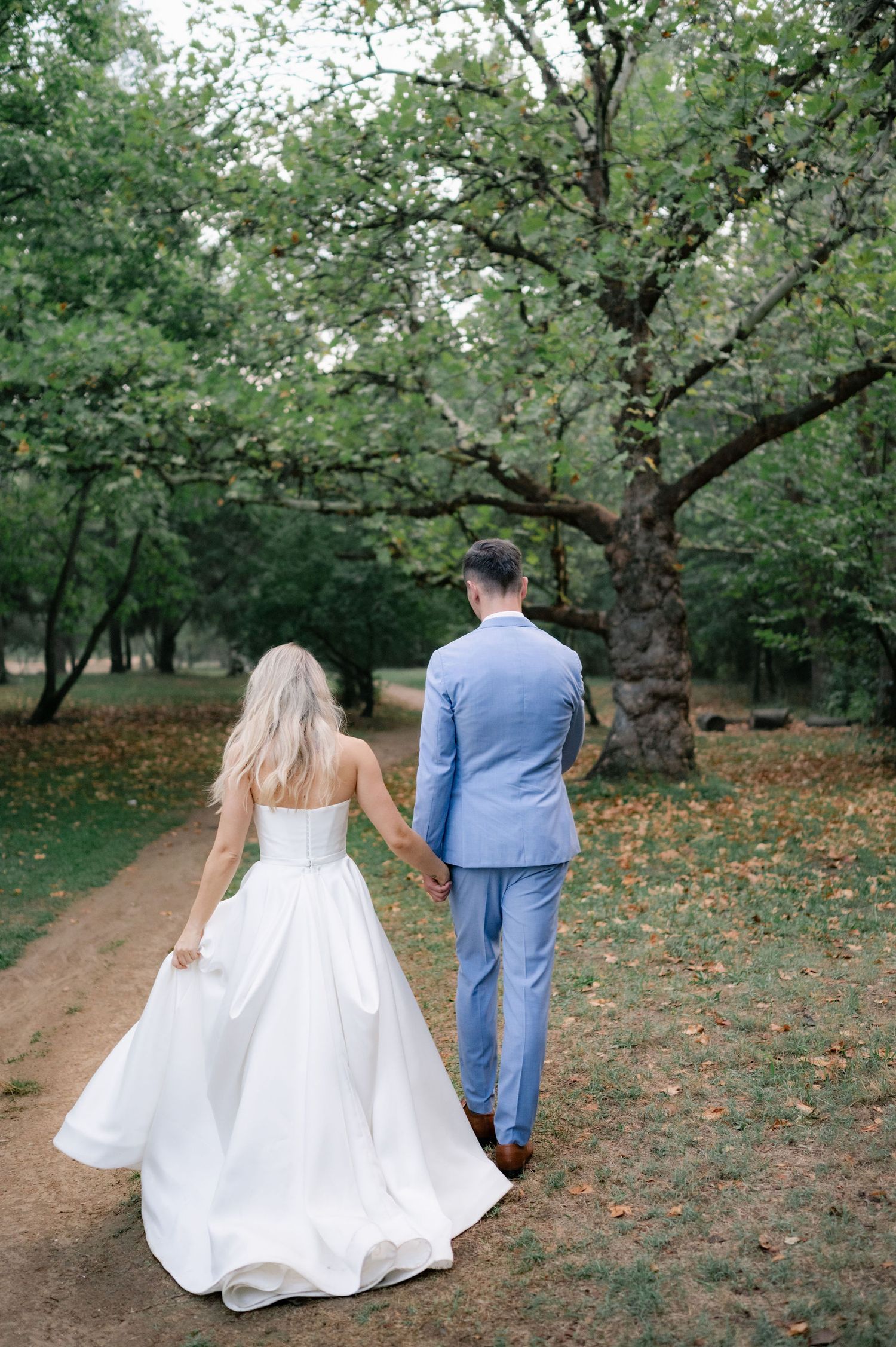 A couple in wedding attire walk hand in hand down a tree-lined dirt path on an autumn day.