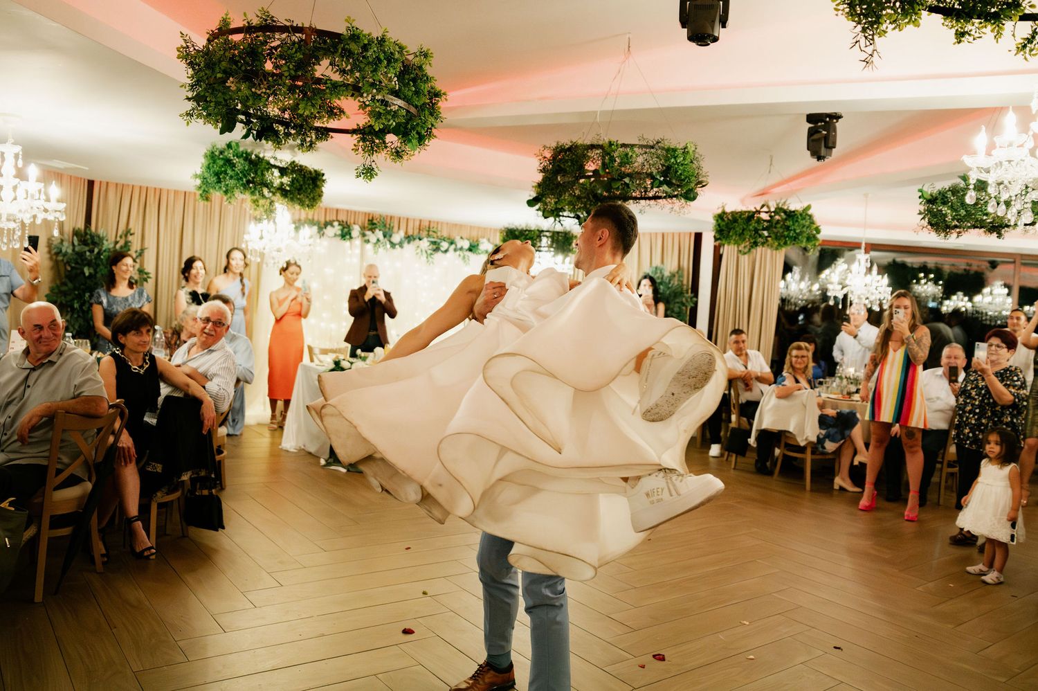 Wedding guests dancing and celebrating on the dance floor under greenery decorations at an indoor reception venue.