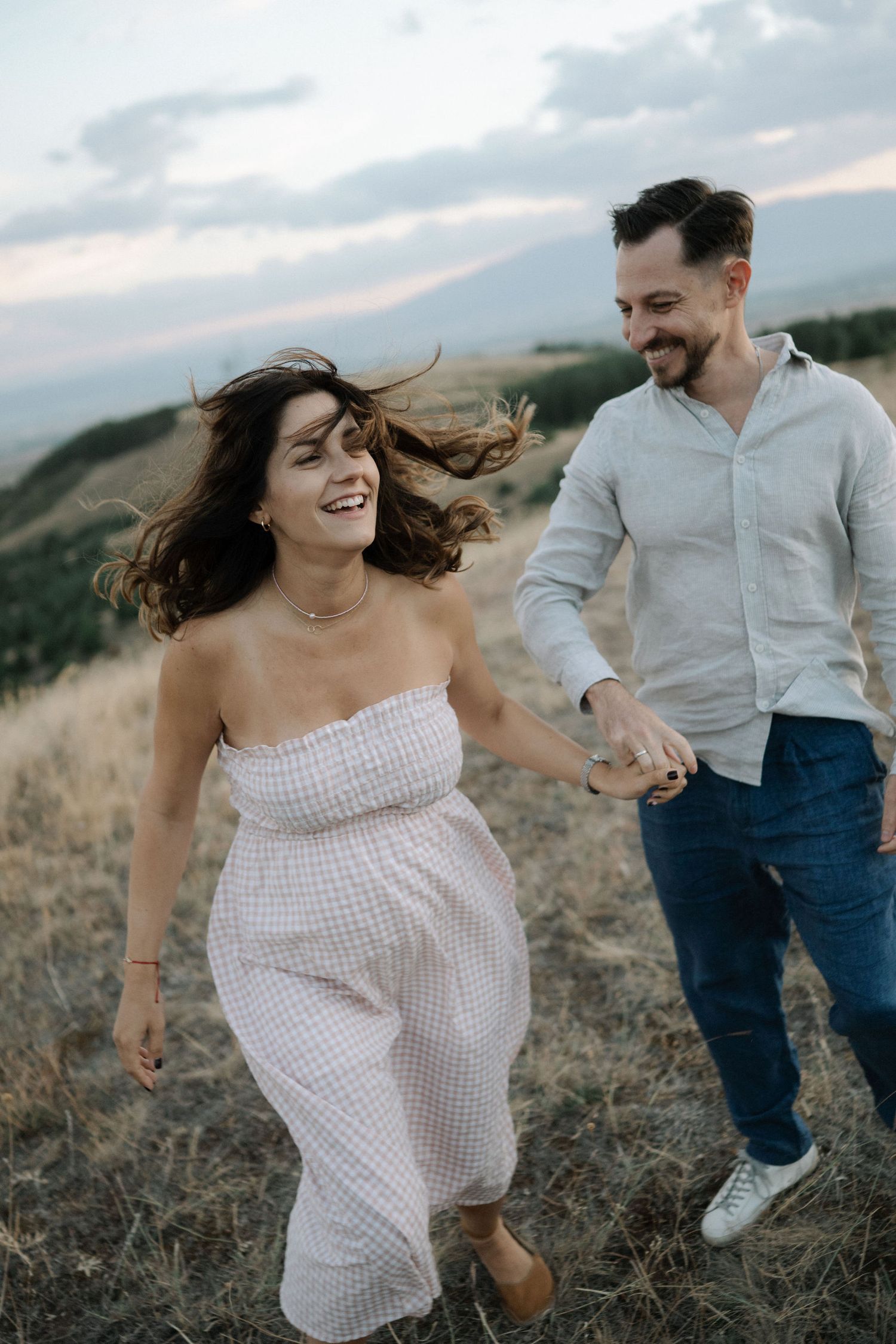 A couple runs playfully through a golden field at sunset, with the person in a white dress laughing joyfully.