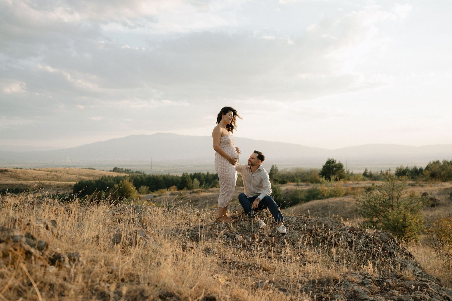Two people sit together on a grassy hilltop overlooking a scenic mountain landscape at sunset.