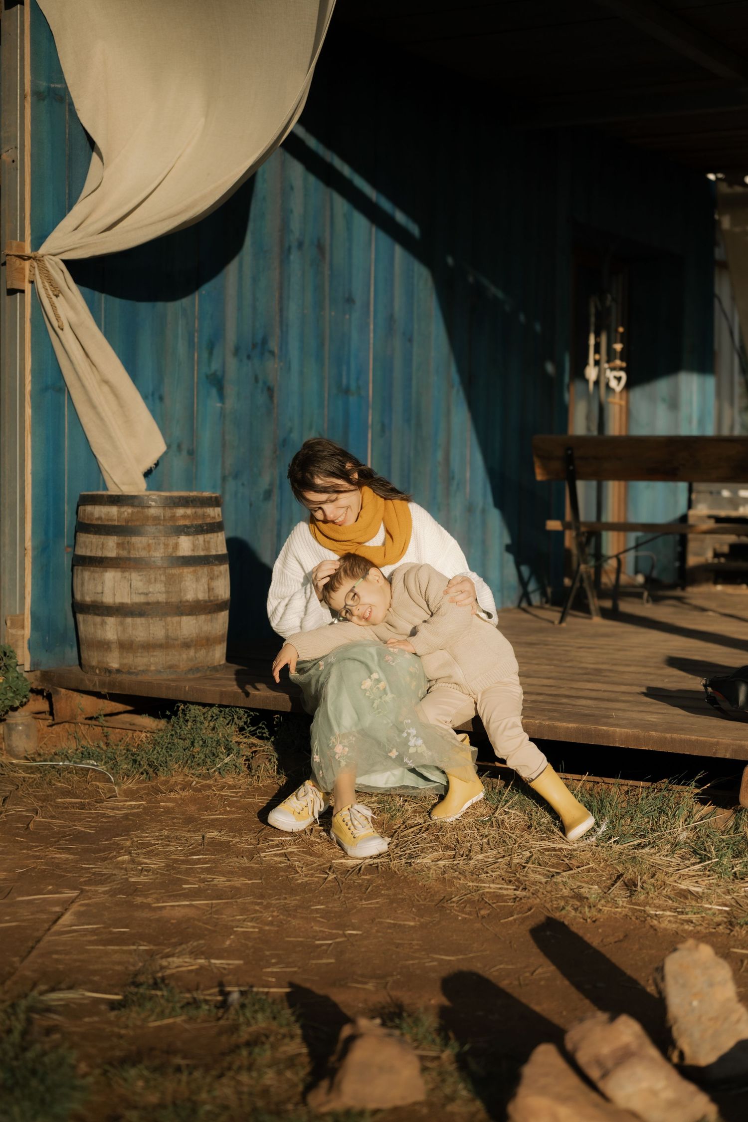 Person in white clothing crouches near wooden barrels and hay on a rustic wooden deck at dusk.