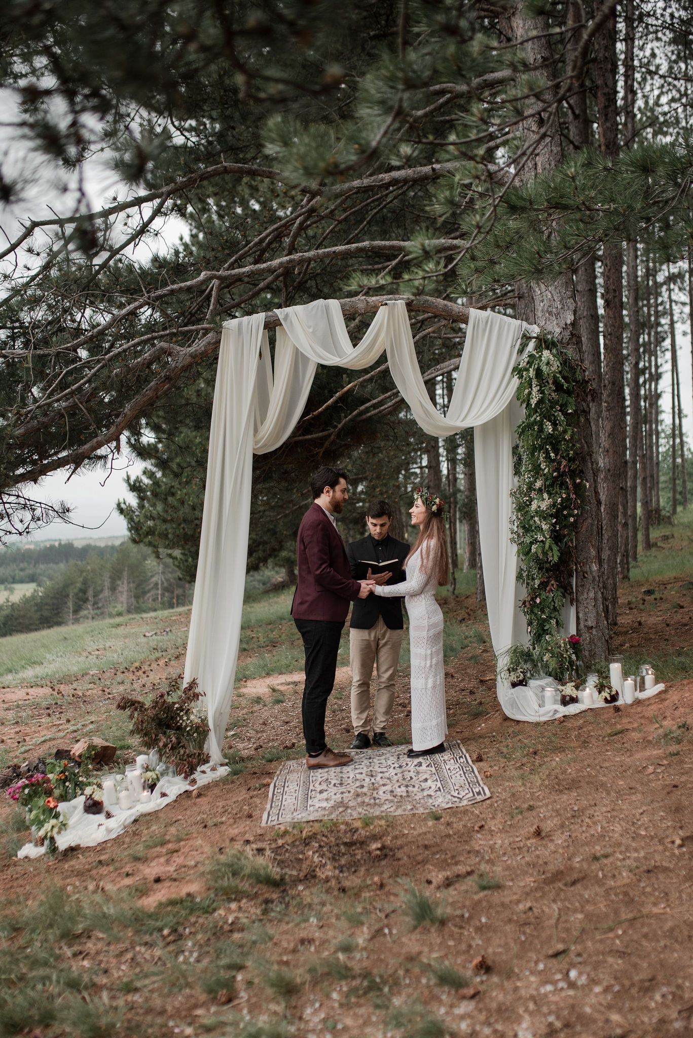 Intimate outdoor wedding ceremony setup in pine forest with flowing white curtains and scattered rose petals on forest floor.