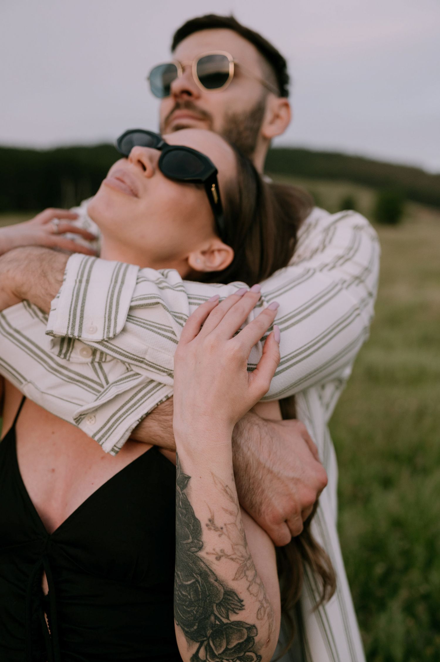 A couple in sunglasses sharing an intimate moment in a grassy field with a focus on their tattoos and casual attire.