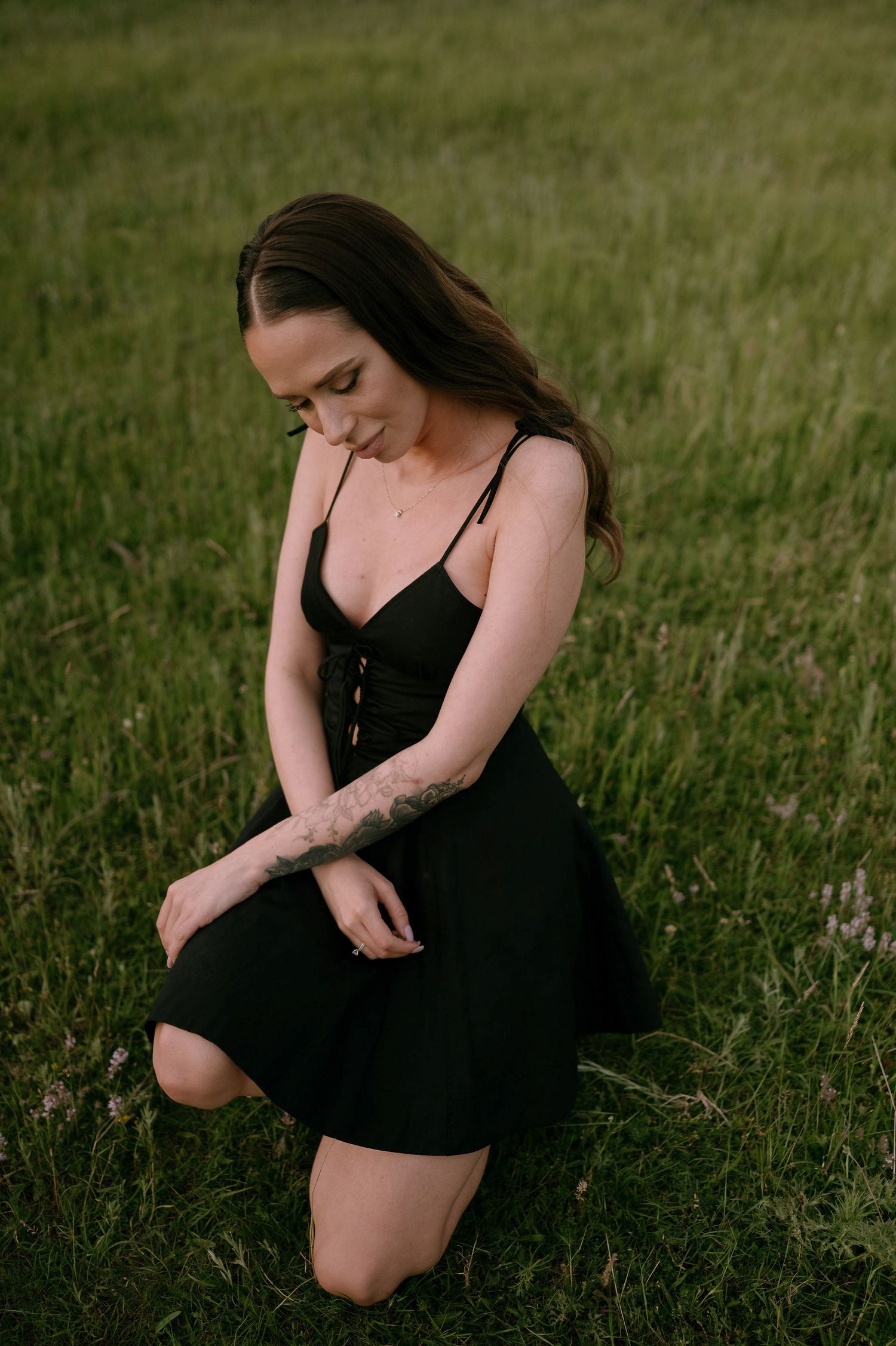A person in a black dress sits thoughtfully in a grassy meadow during golden hour.