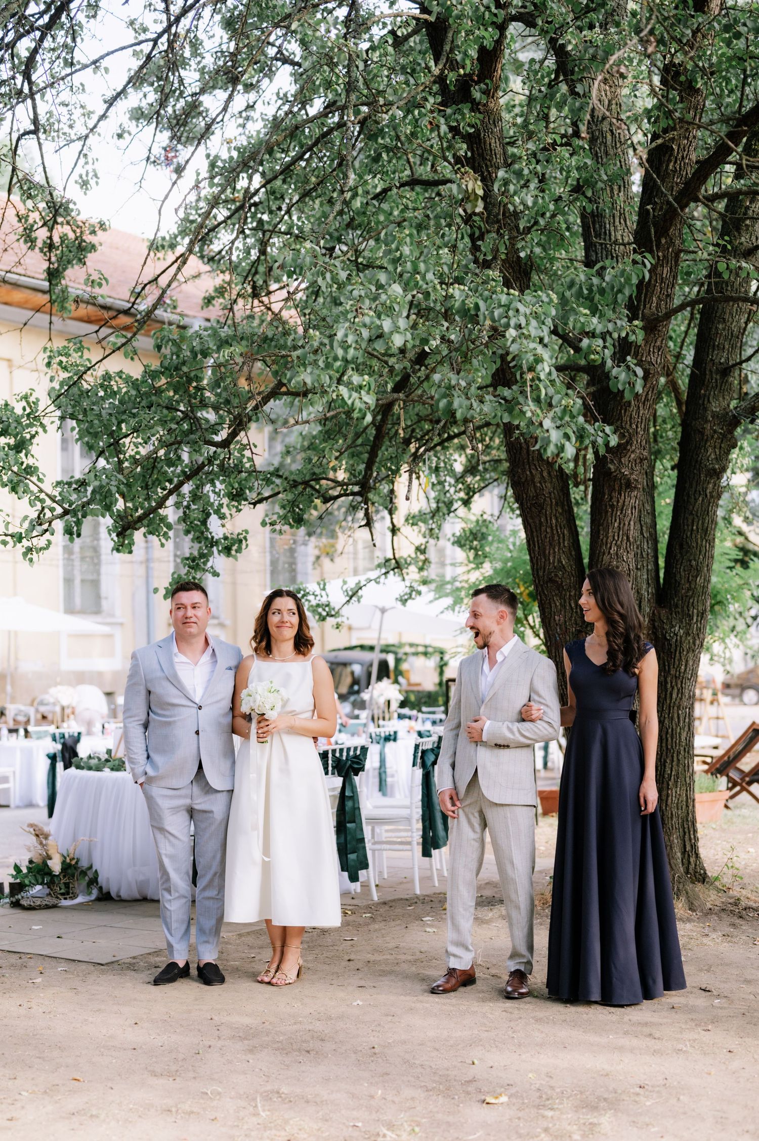 Elegant wedding guests in formal attire gather under a large tree at an outdoor summer celebration.