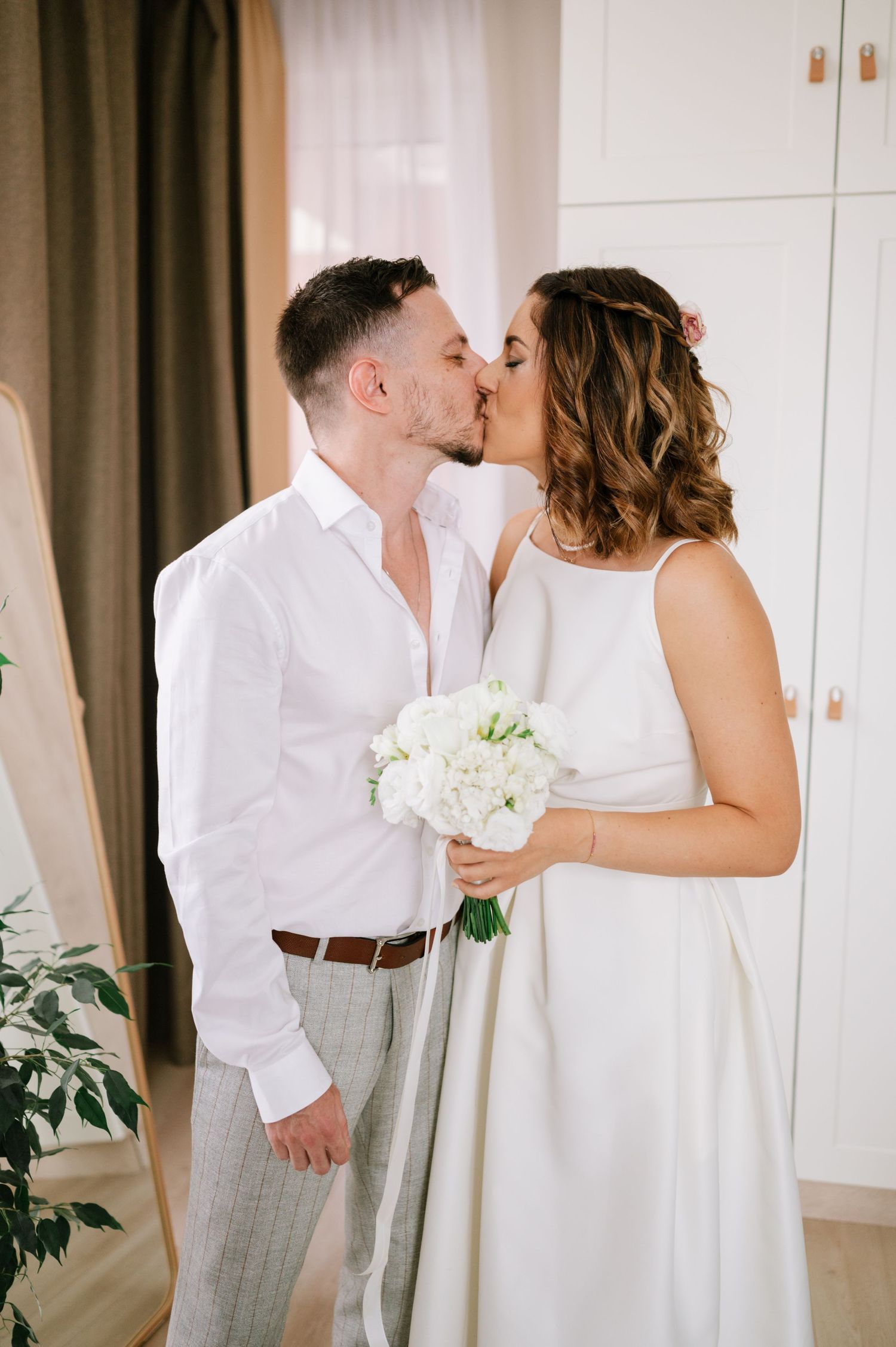 A couple in white wedding attire share a romantic kiss while holding a bouquet of white flowers.