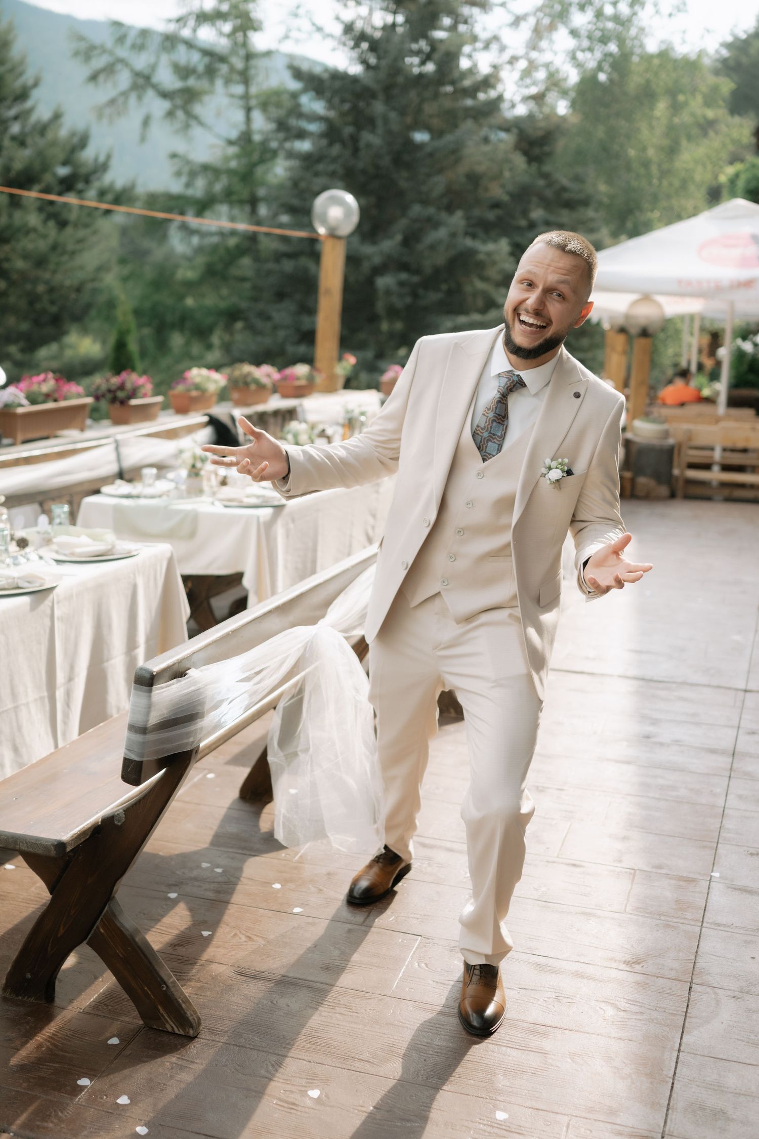 A person in a cream-colored three-piece suit dances joyfully at an outdoor wedding reception with tables in the background.