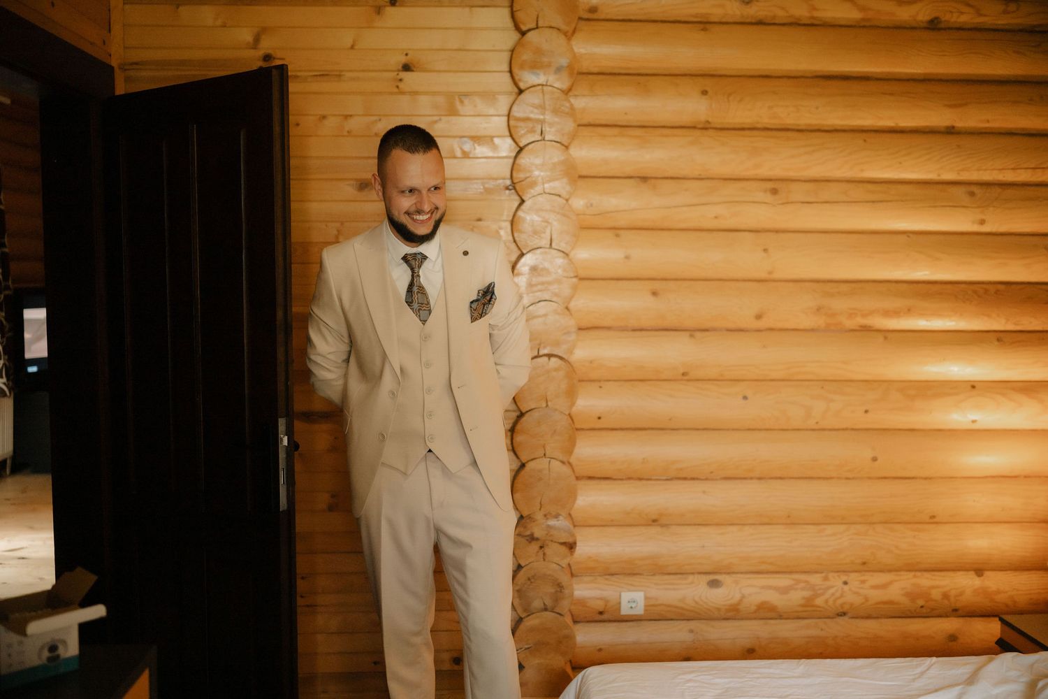 Someone in a light beige suit stands against rustic wooden log cabin walls.