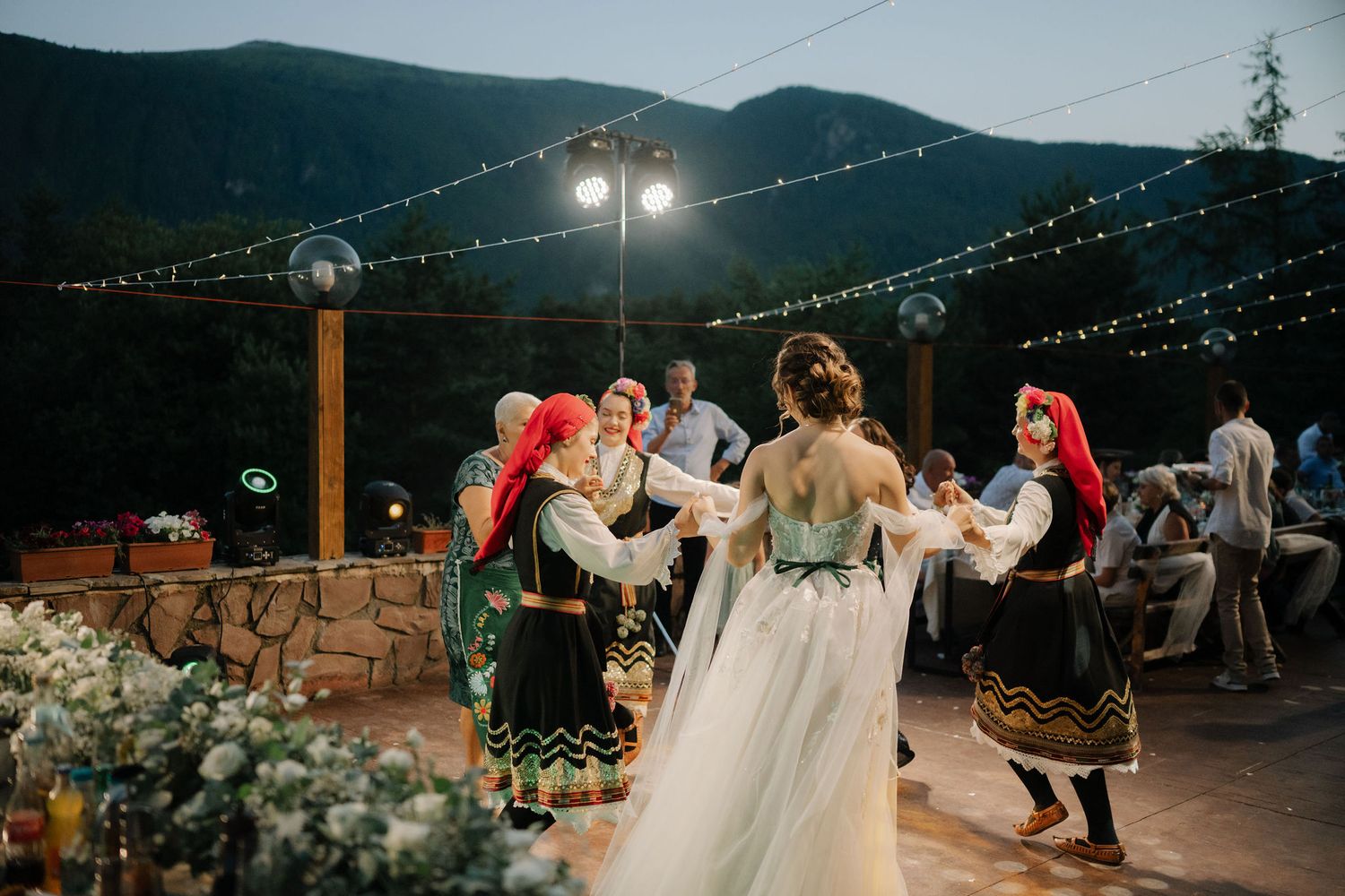 People dance during an outdoor evening wedding reception with string lights and mountain views in the background.