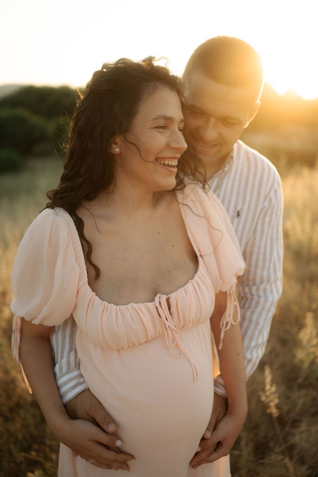 A couple shares an intimate moment in a sunlit field during a maternity photoshoot at golden hour.