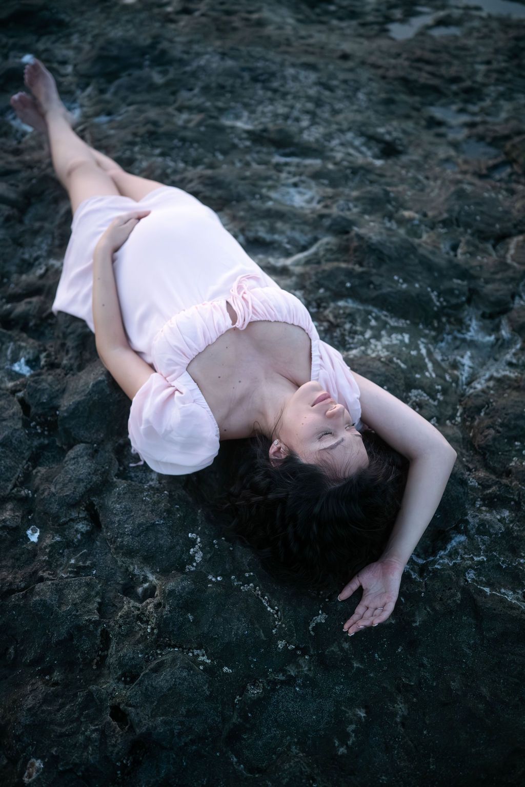 Person in pale dress reclines on dark stone surface in moody atmospheric setting.