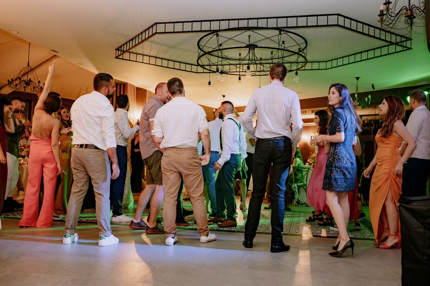 Wedding guests dance together on an illuminated dance floor under decorative ceiling lighting at an indoor reception.