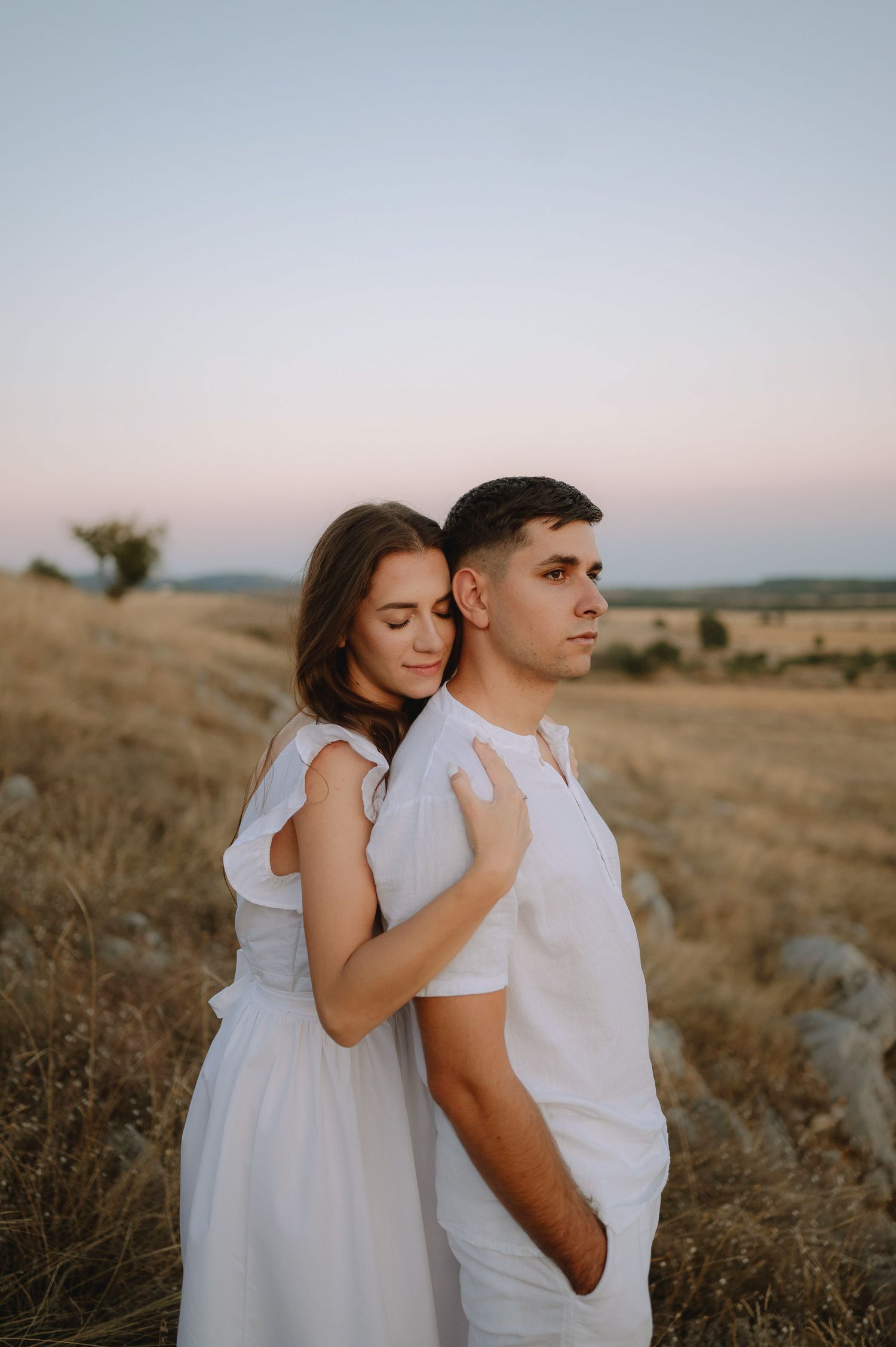 Couple in white outfits embrace tenderly in a golden field at sunset creating a romantic outdoor portrait.