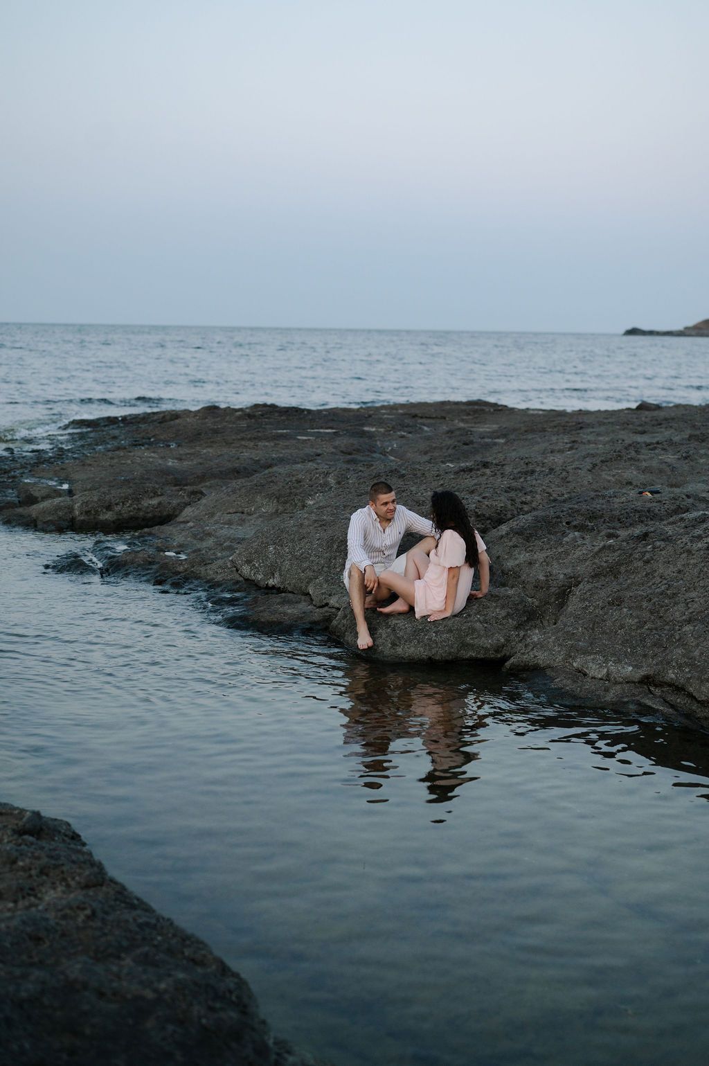 A couple sits together on volcanic rocks by the ocean at dusk, creating a romantic coastal scene.