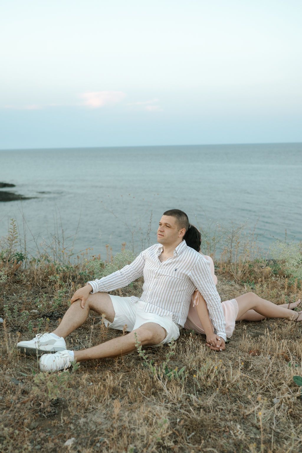 A couple relaxes together on a coastal cliff with the sea and horizon in the background.