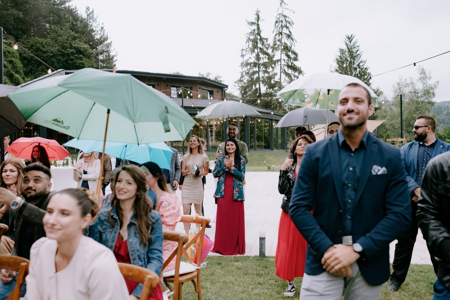 Wedding guests huddle under umbrellas during an outdoor ceremony on a rainy day.