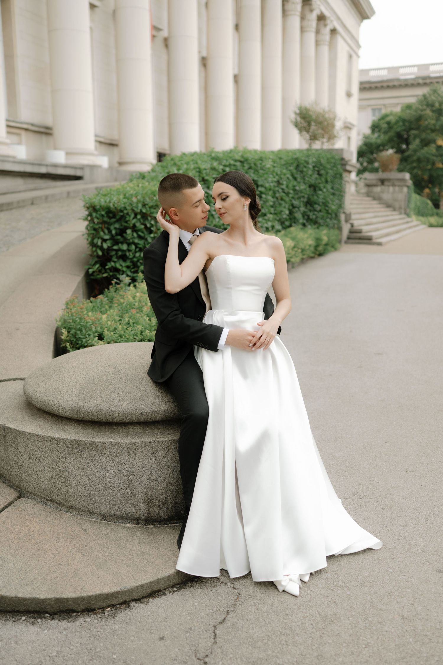 A couple in wedding attire pose elegantly on stone steps against a classical building with white columns.