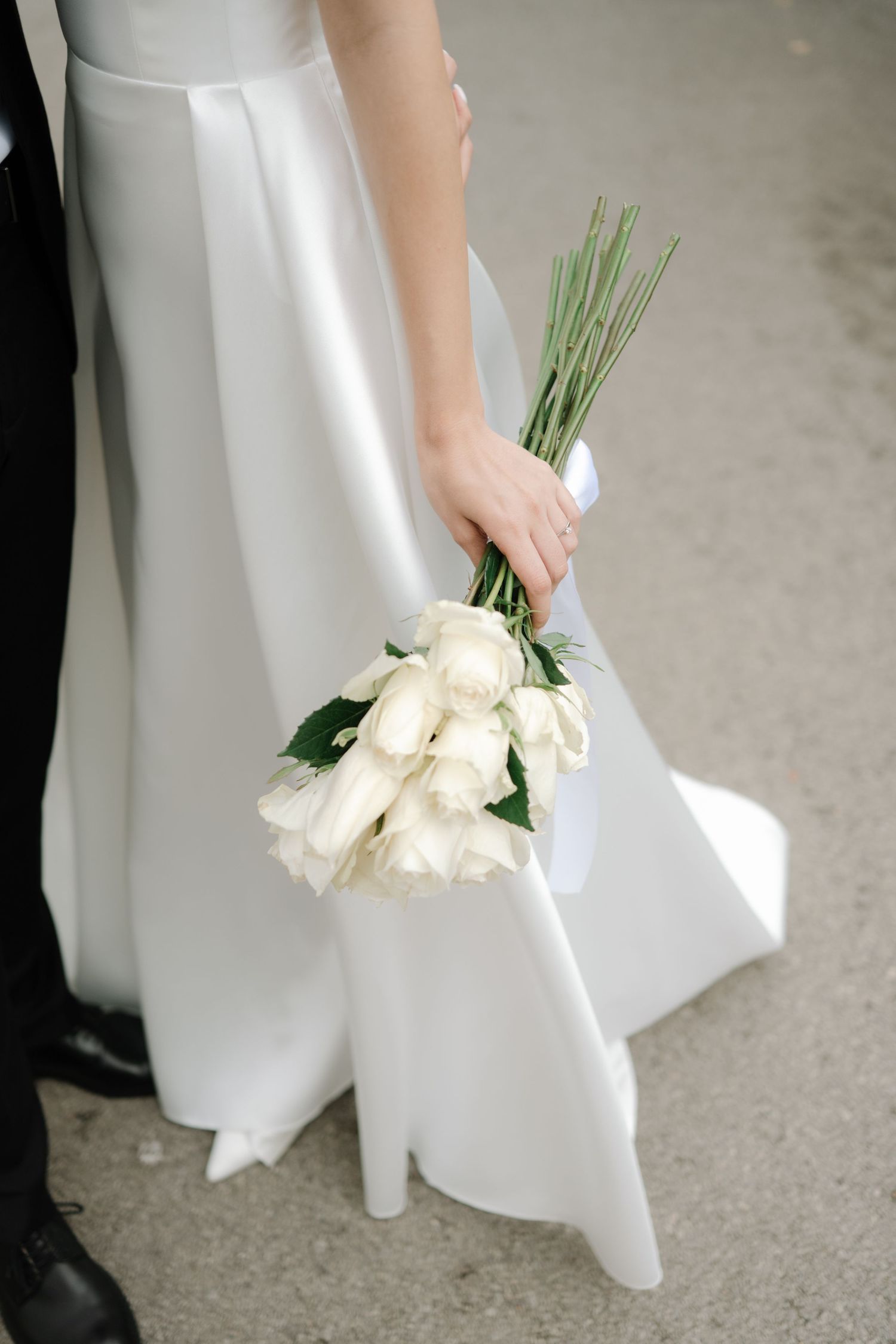 A bride in a white wedding gown holds a classic bouquet of white roses against a grey background.