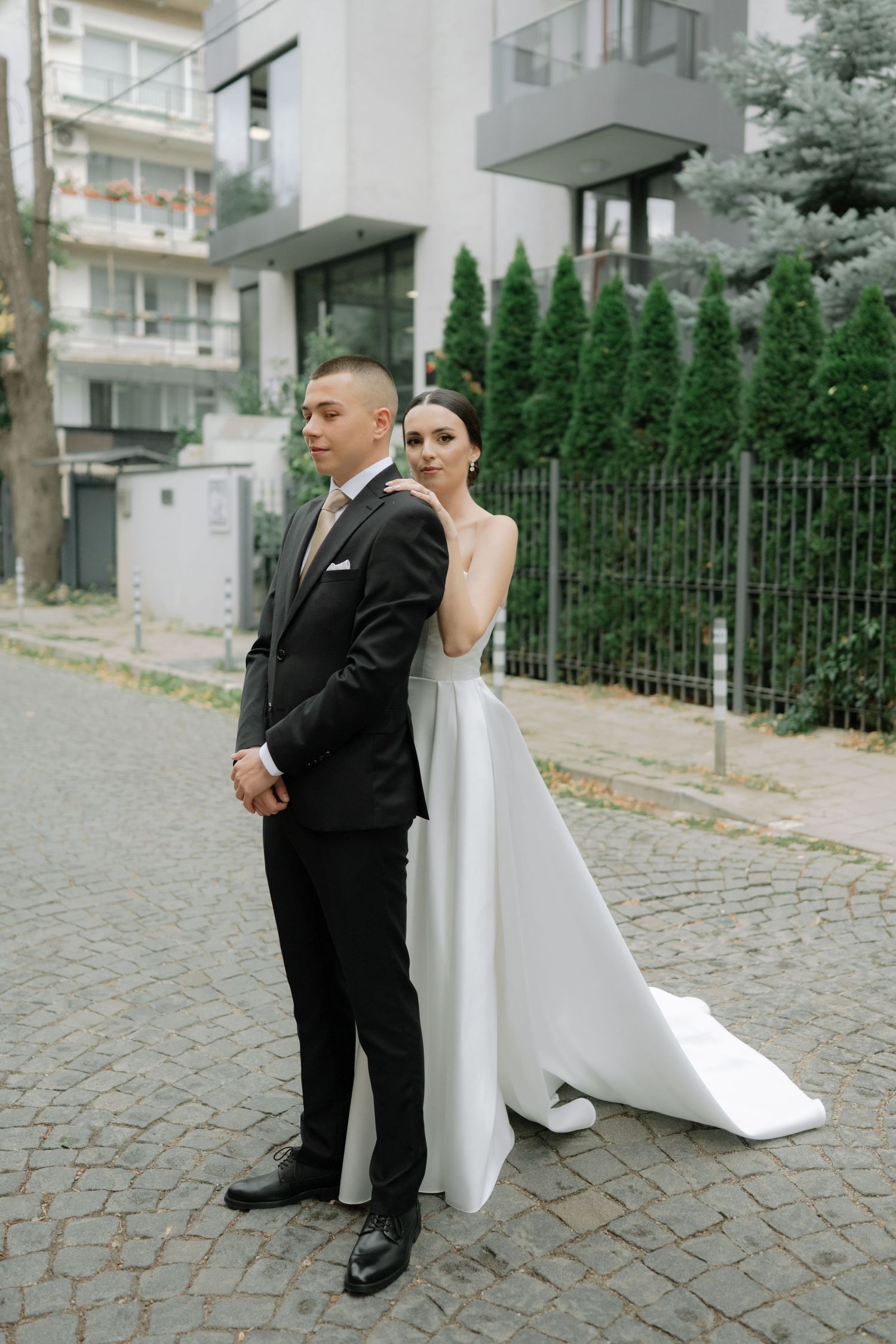 A couple poses back-to-back on a cobblestone street for a wedding photo with modern apartments and evergreens in background.