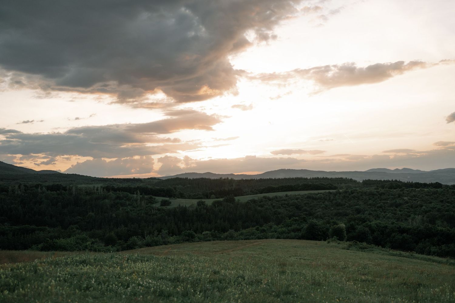 Scenic sunset over rolling green hills and forest landscape with dramatic cloudy sky in a rural countryside setting.