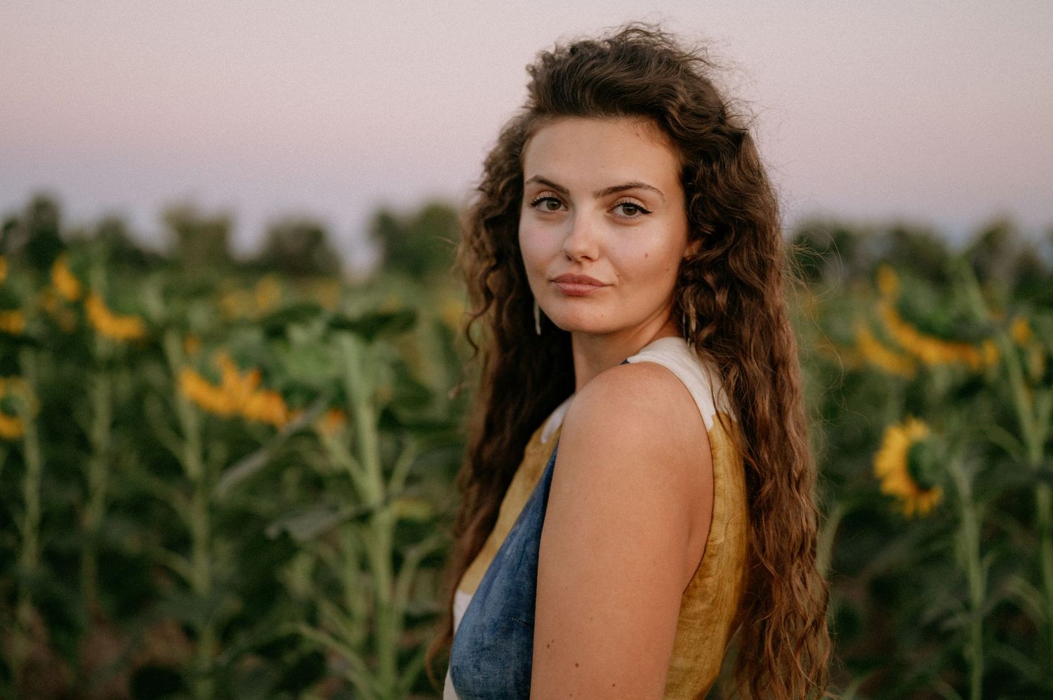 Young woman with curly hair posing in a sunflower field at sunset, wearing a denim top.