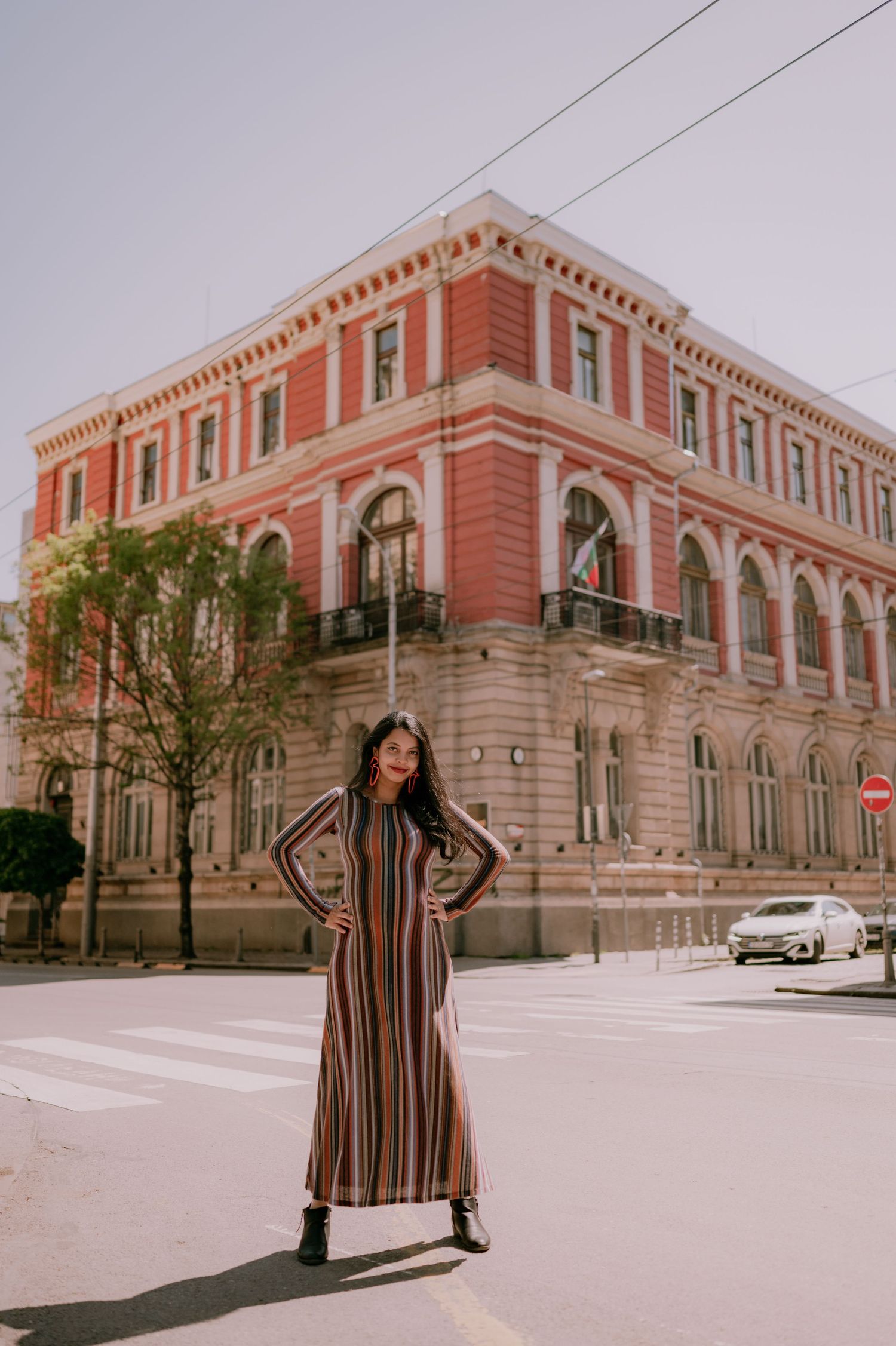 Woman in striped maxi dress posing confidently in front of a grand red historic building on a sunny day.