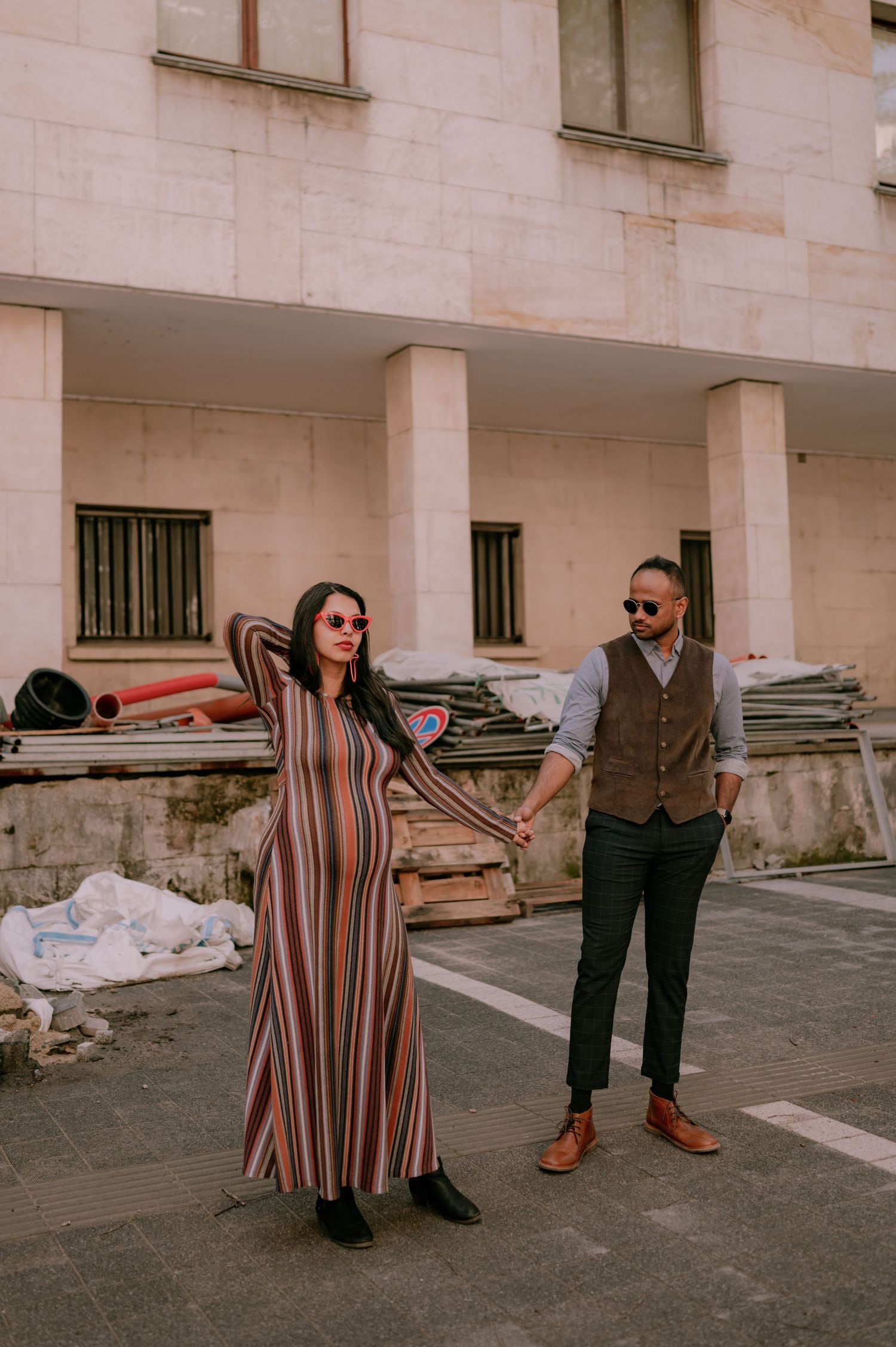 Stylish couple holding hands outdoors, woman in striped maxi dress and man in vest and trousers, urban building backdrop.