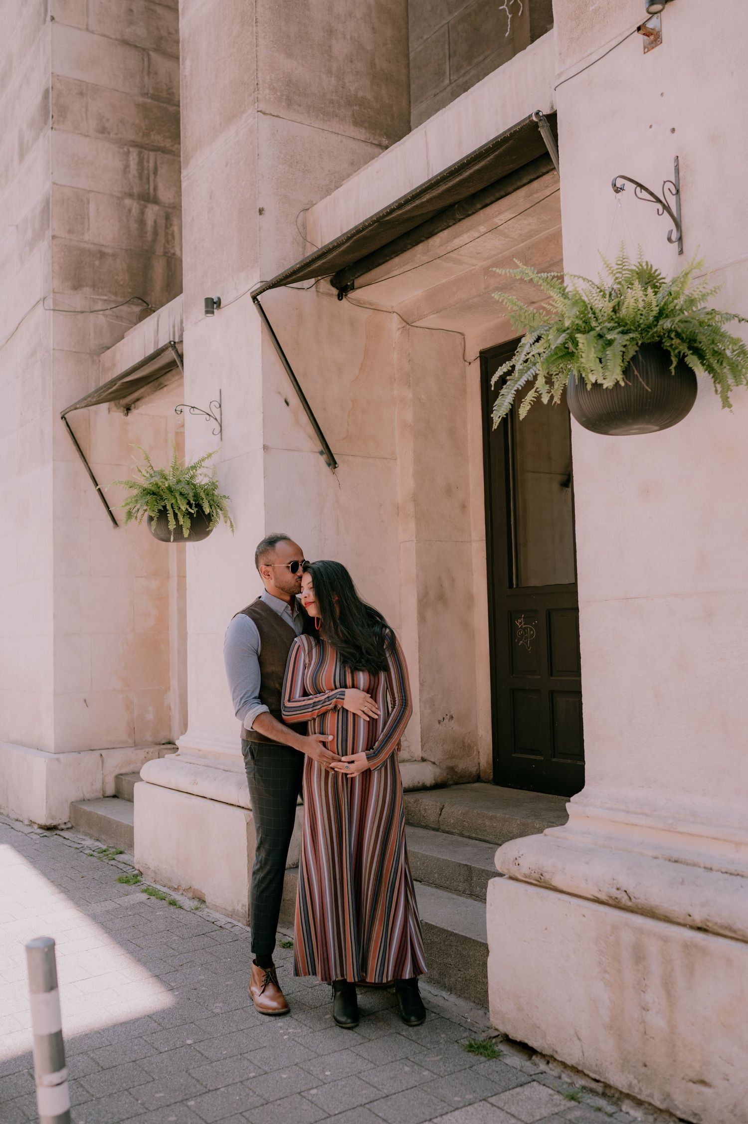 Couple sharing a tender moment outside a stone building with hanging ferns, woman in striped maxi dress cradling baby bump.