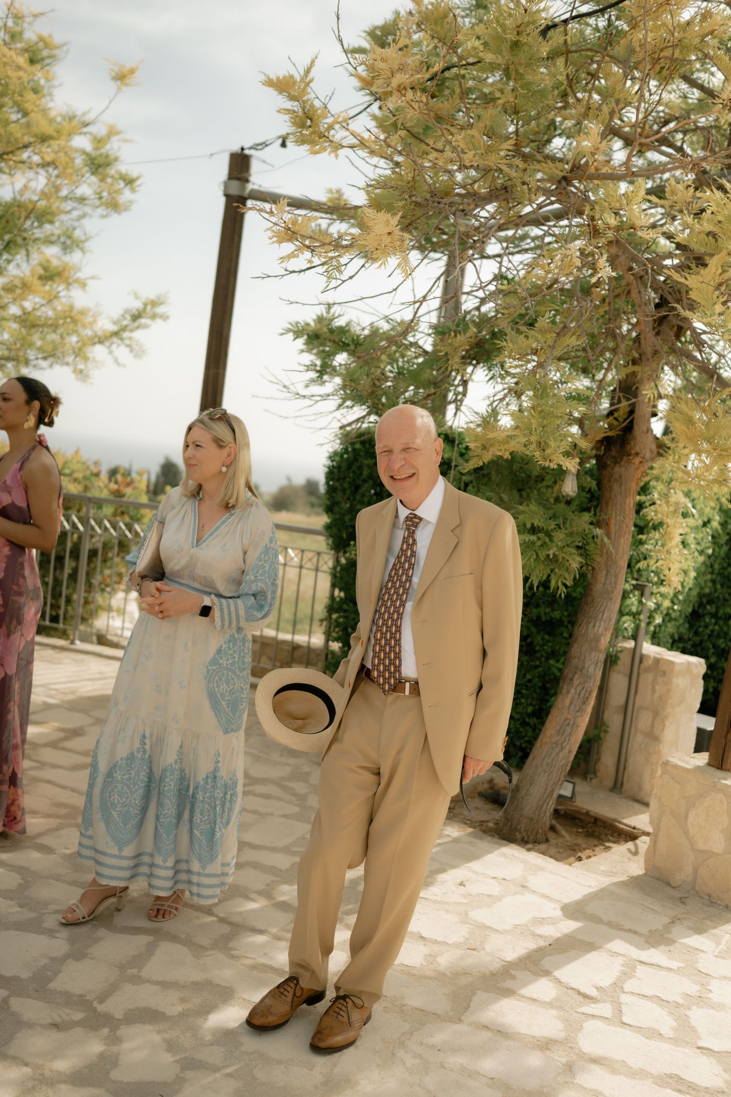 Elegant couple in beige suit and floral dress at outdoor wedding venue with stone patio and trees.