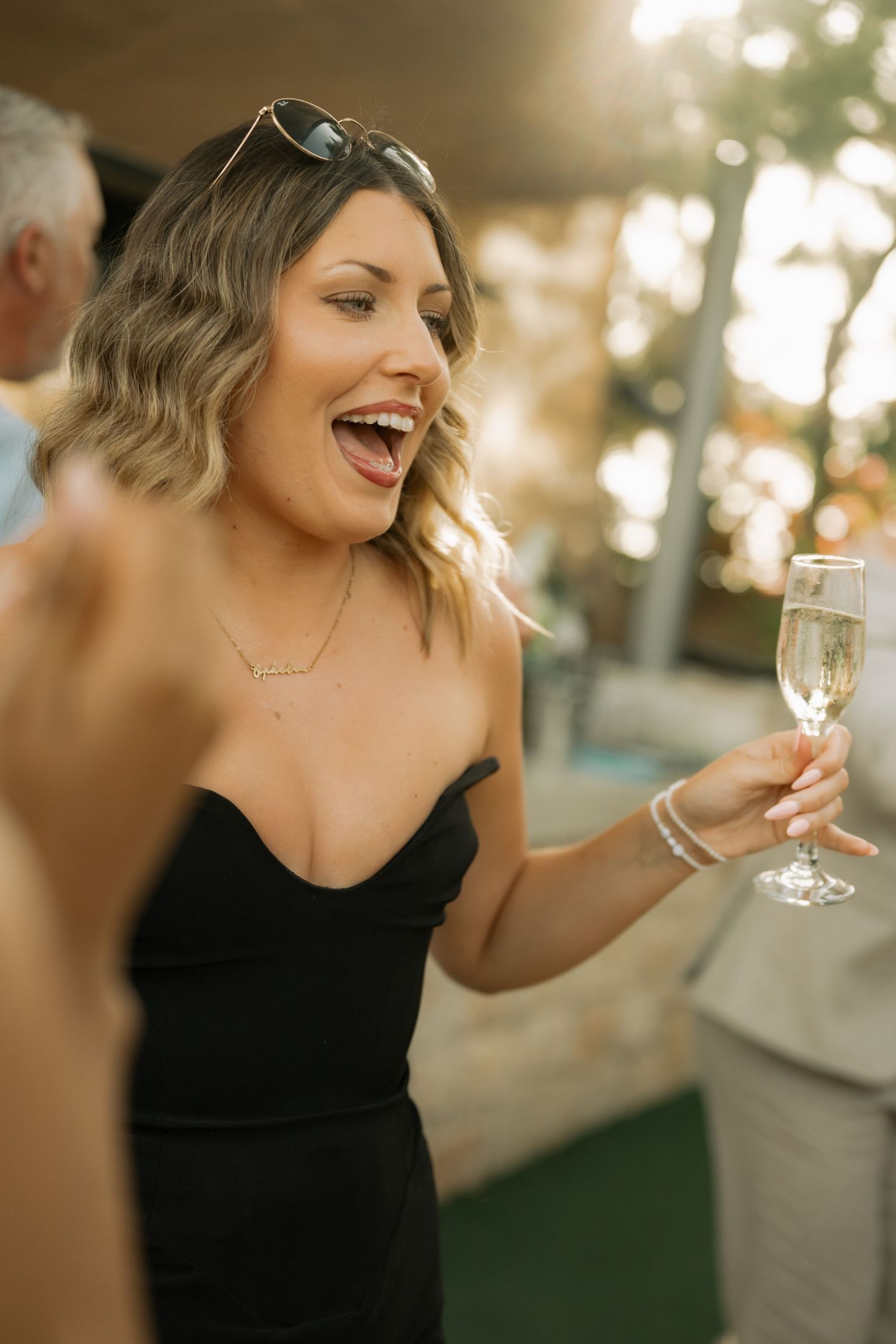 Smiling woman in black dress holding champagne flute at outdoor party in golden sunset light.