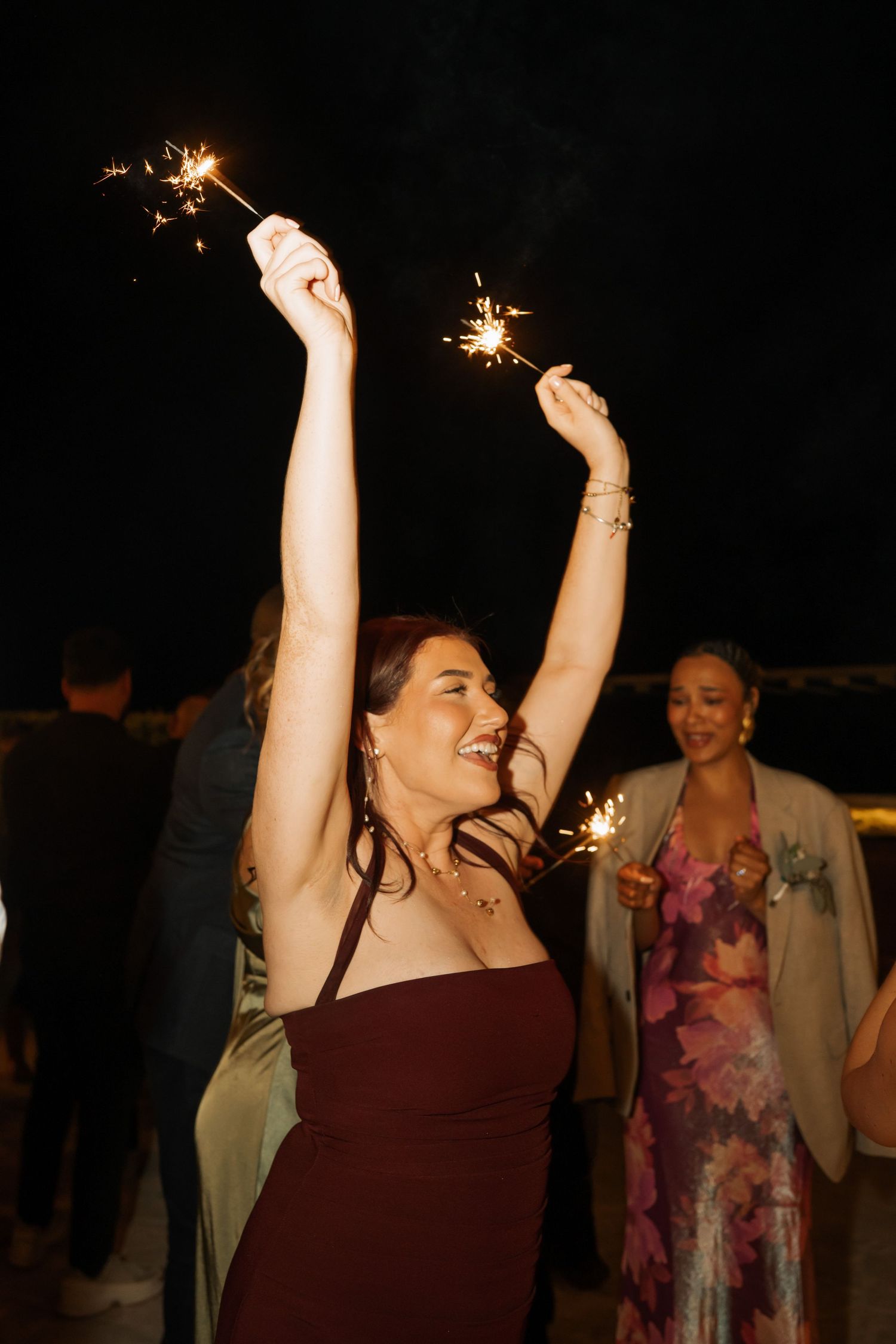 Woman in burgundy dress joyfully holds sparklers at a nighttime wedding celebration with guests in background.