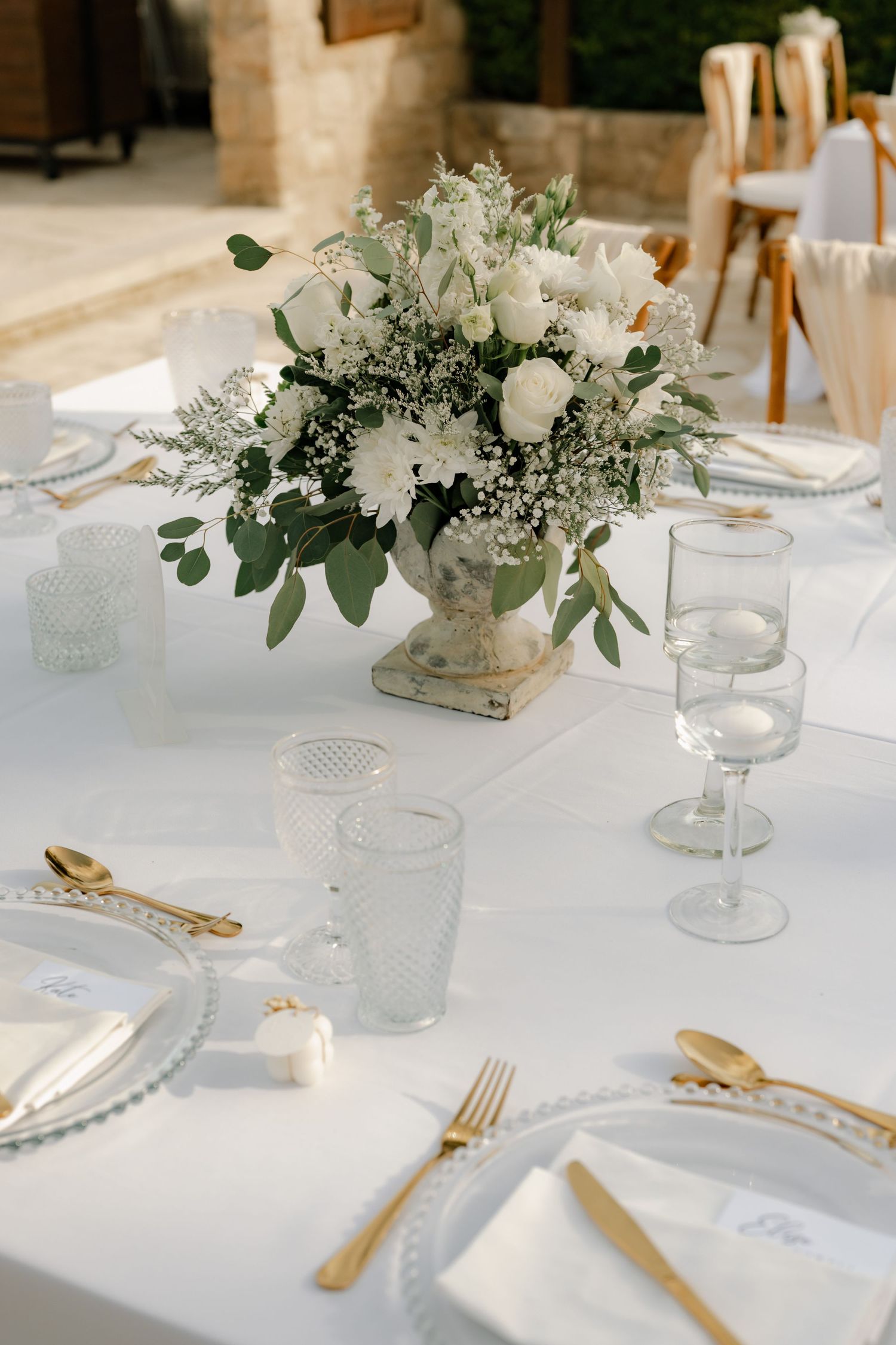 Elegant wedding table setting with white floral centerpiece, gold cutlery, crystal glasses, and white linen tablecloth.