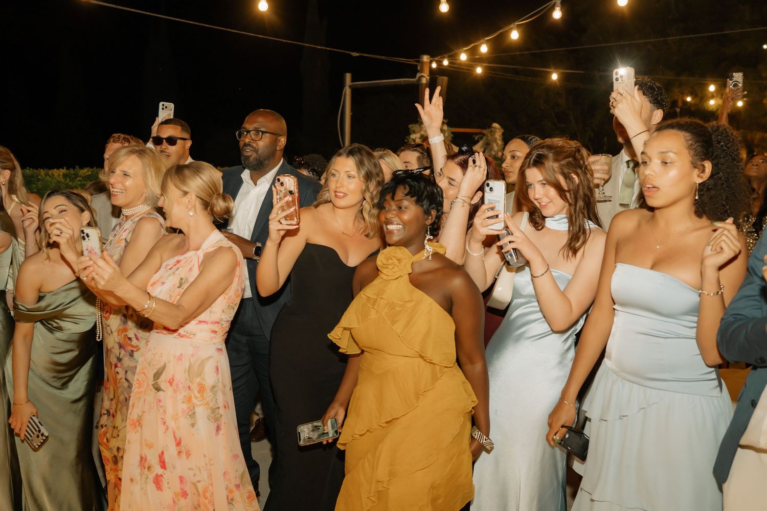 Diverse wedding guests dancing and taking photos at an outdoor evening reception under string lights.