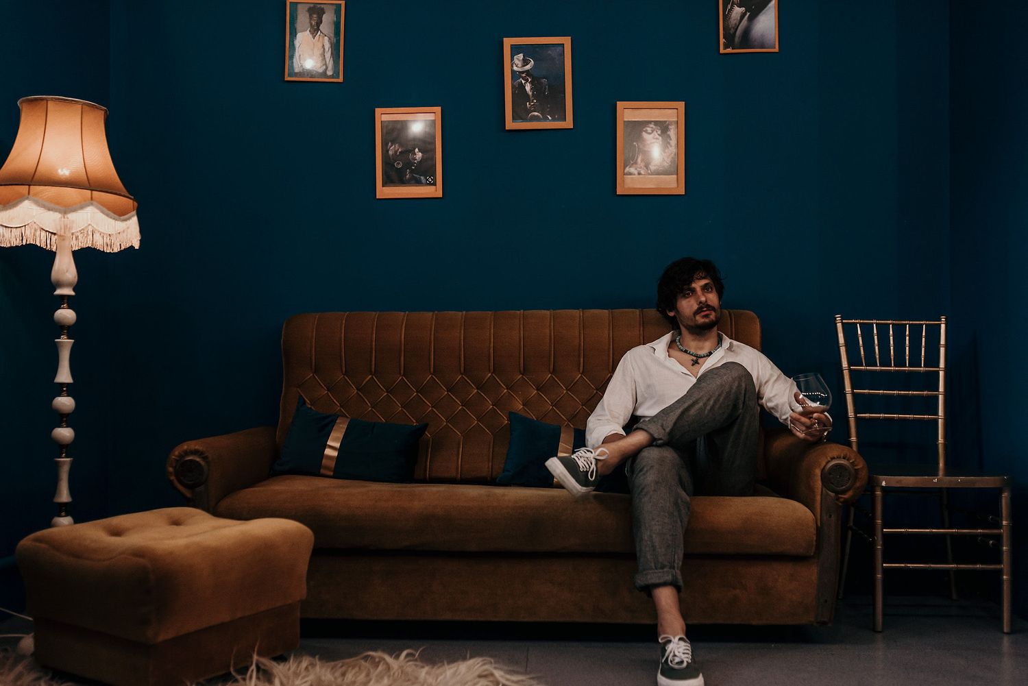 Person relaxing on brown leather couch with vintage floor lamp and photo wall.