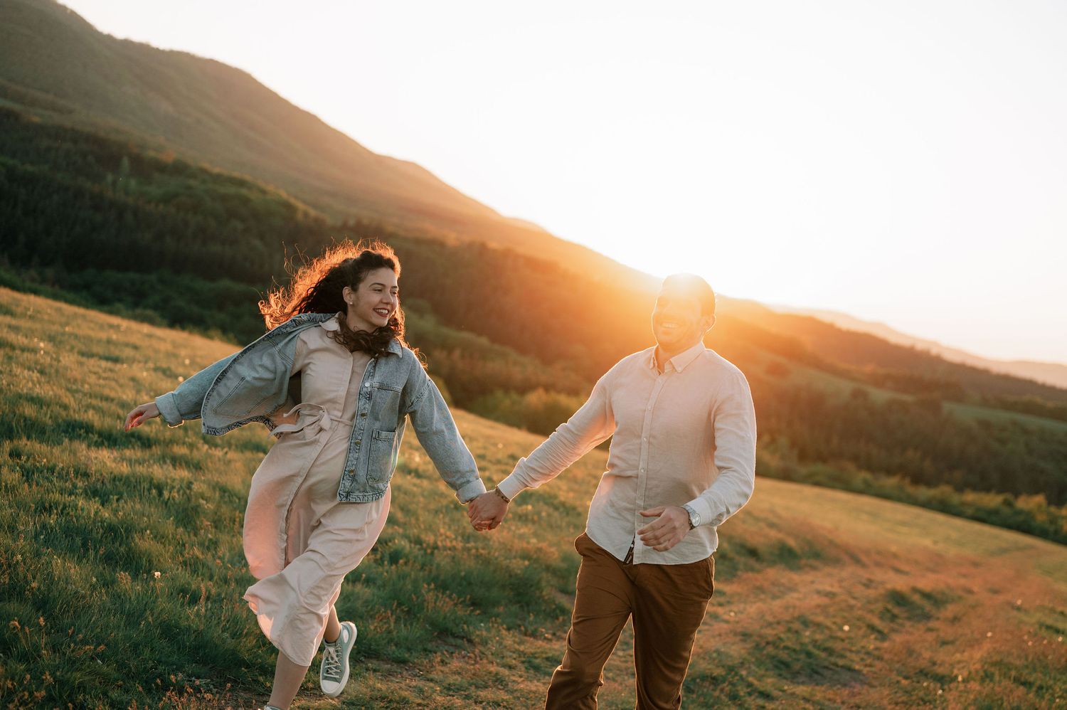 Couple holding hands and running through a grassy hillside during golden hour sunset.
