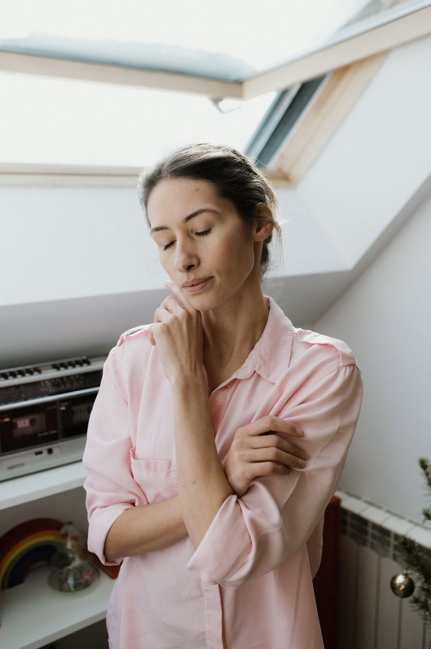 Person in pink button-up shirt stands thoughtfully near window with eyes closed in natural daylight.