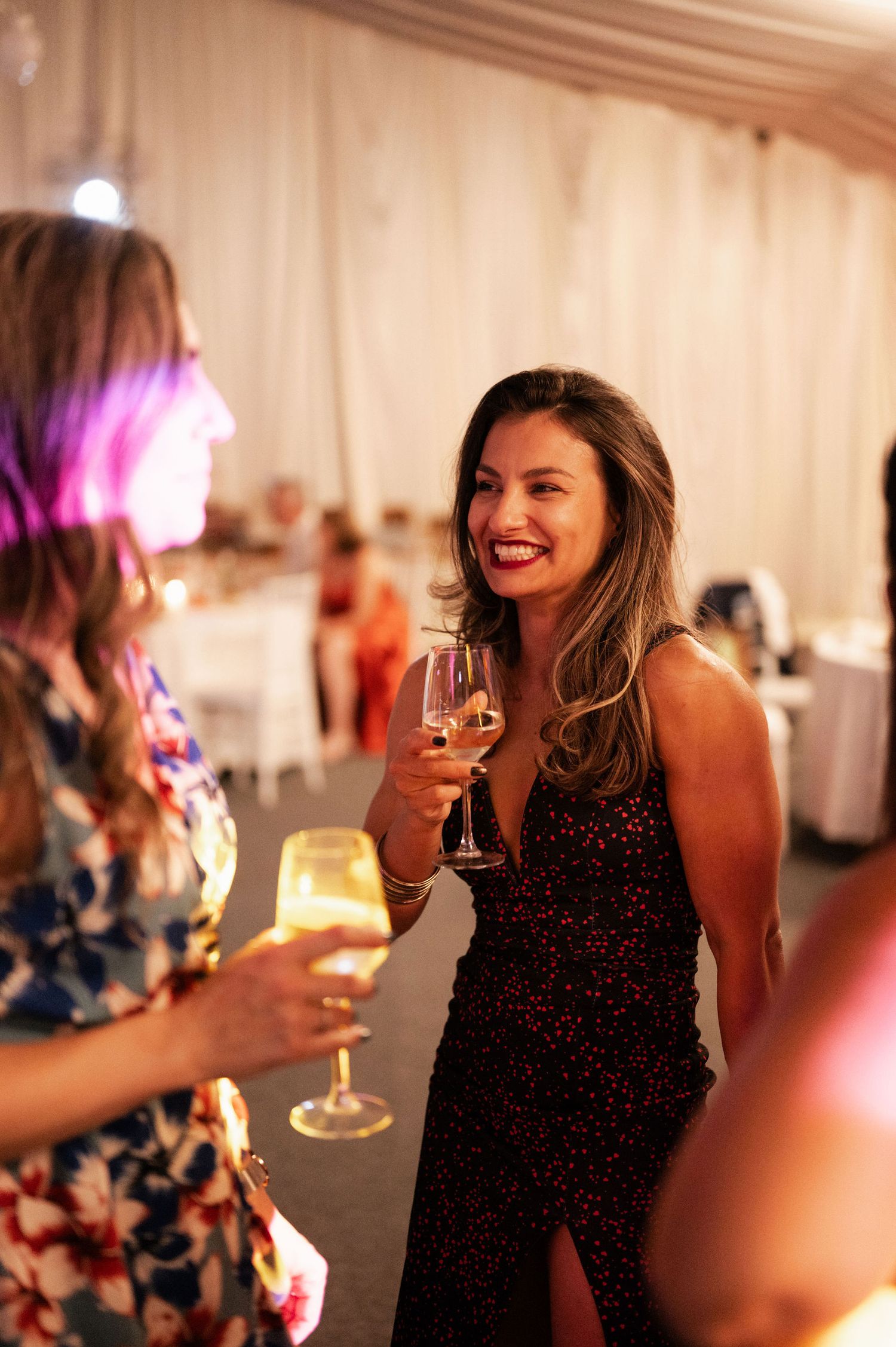 A person in a black sparkly dress laughs while holding a wine glass at an elegant indoor event with soft lighting.