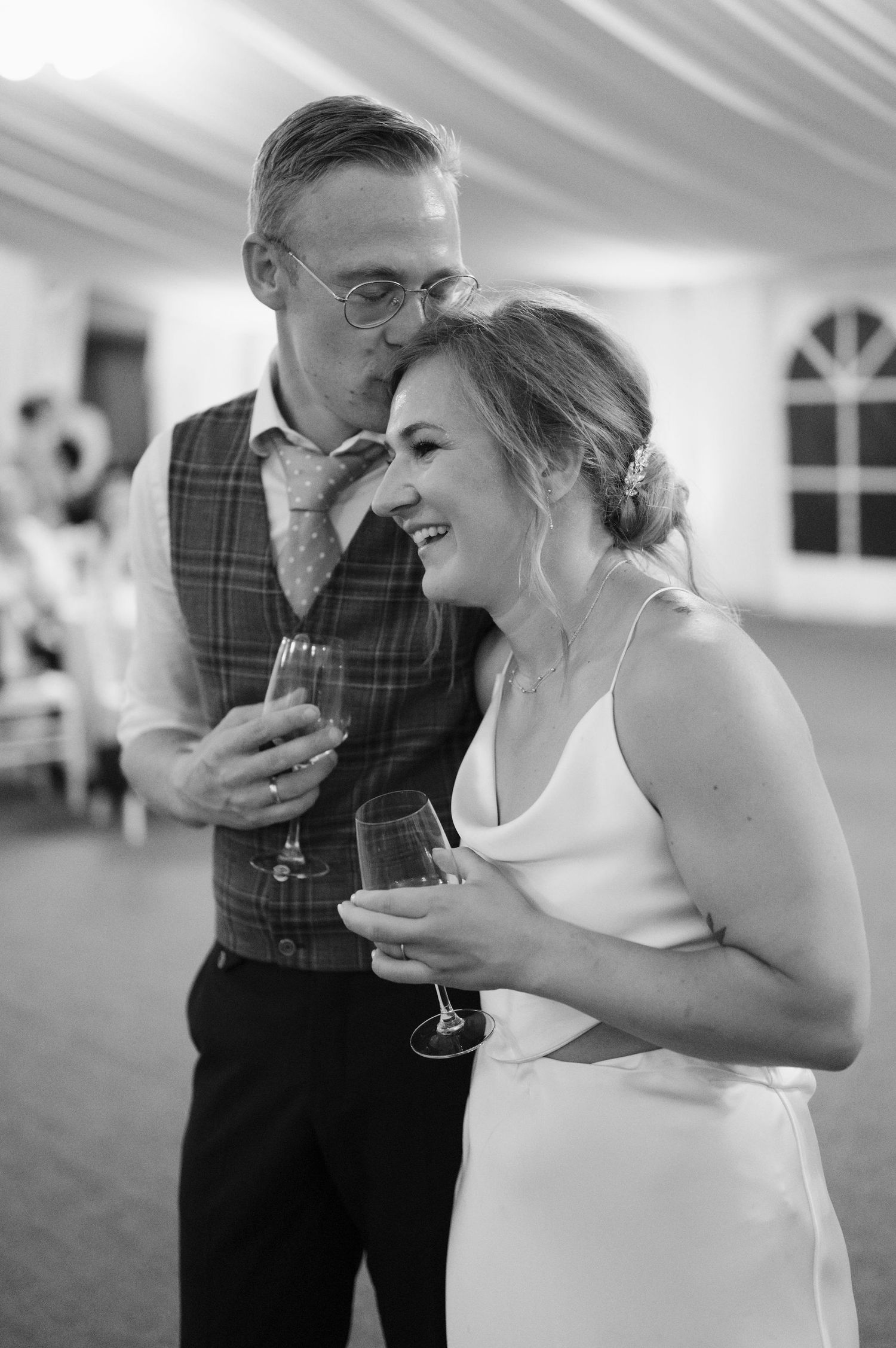 Black and white candid photo of a couple laughing together at a wedding reception while holding drinks.