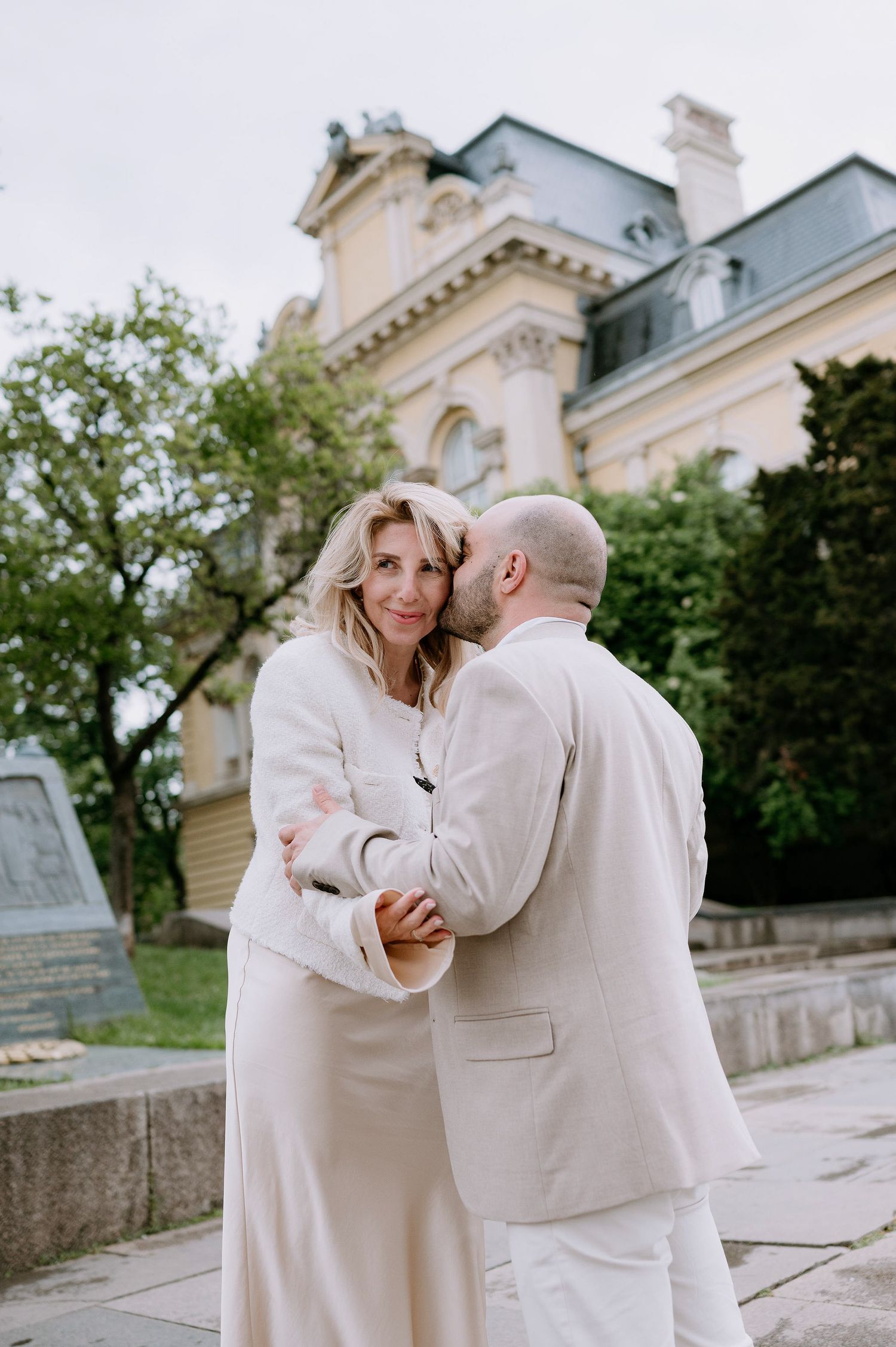A couple dressed in cream-colored outfits shares a romantic embrace near a classical building.