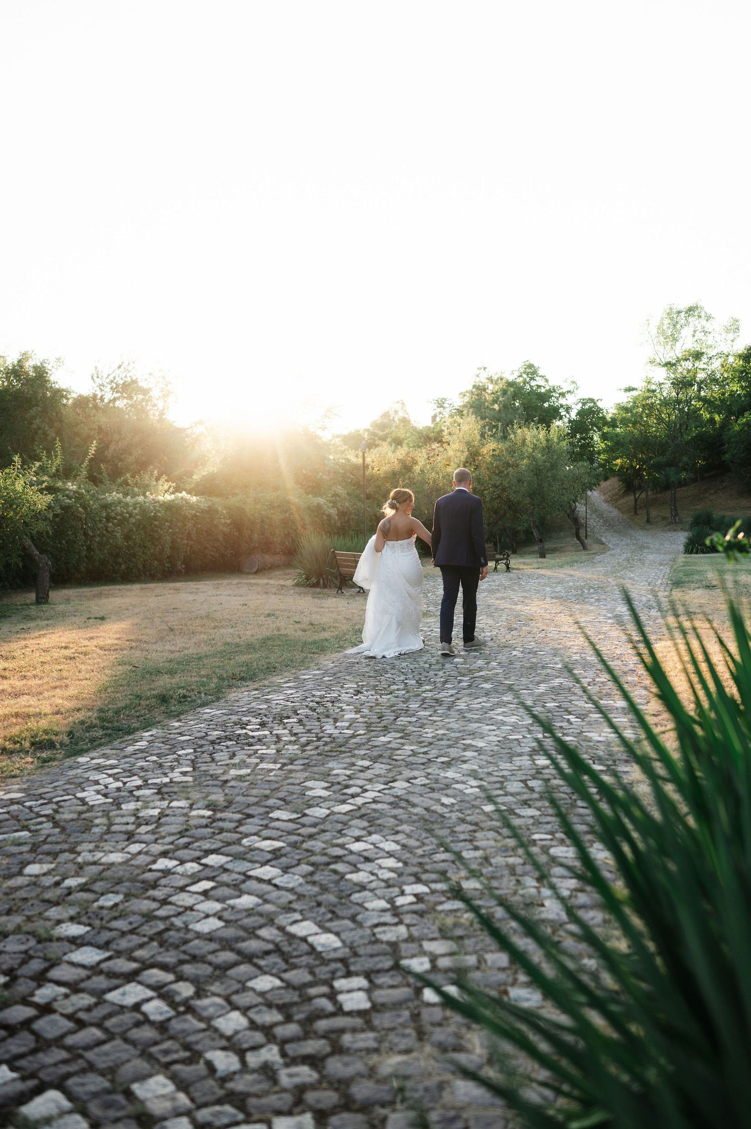 Couple walking along a sunlit cobblestone path with lush greenery in the background.