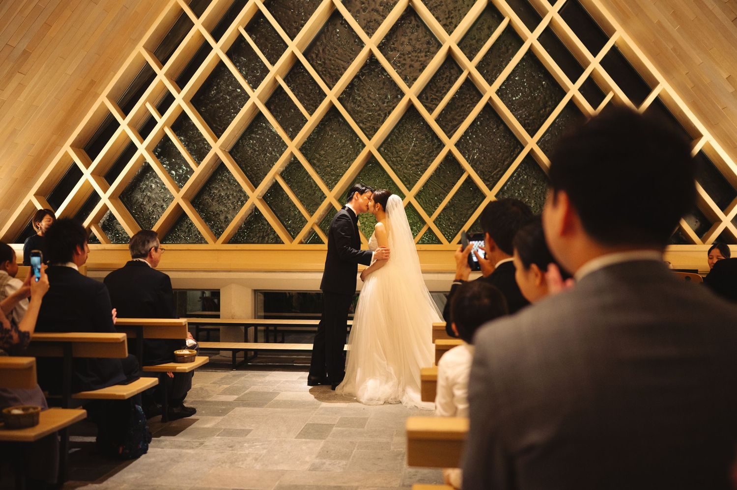 Wedding ceremony taking place in a contemporary chapel with geometric wooden wall patterns.