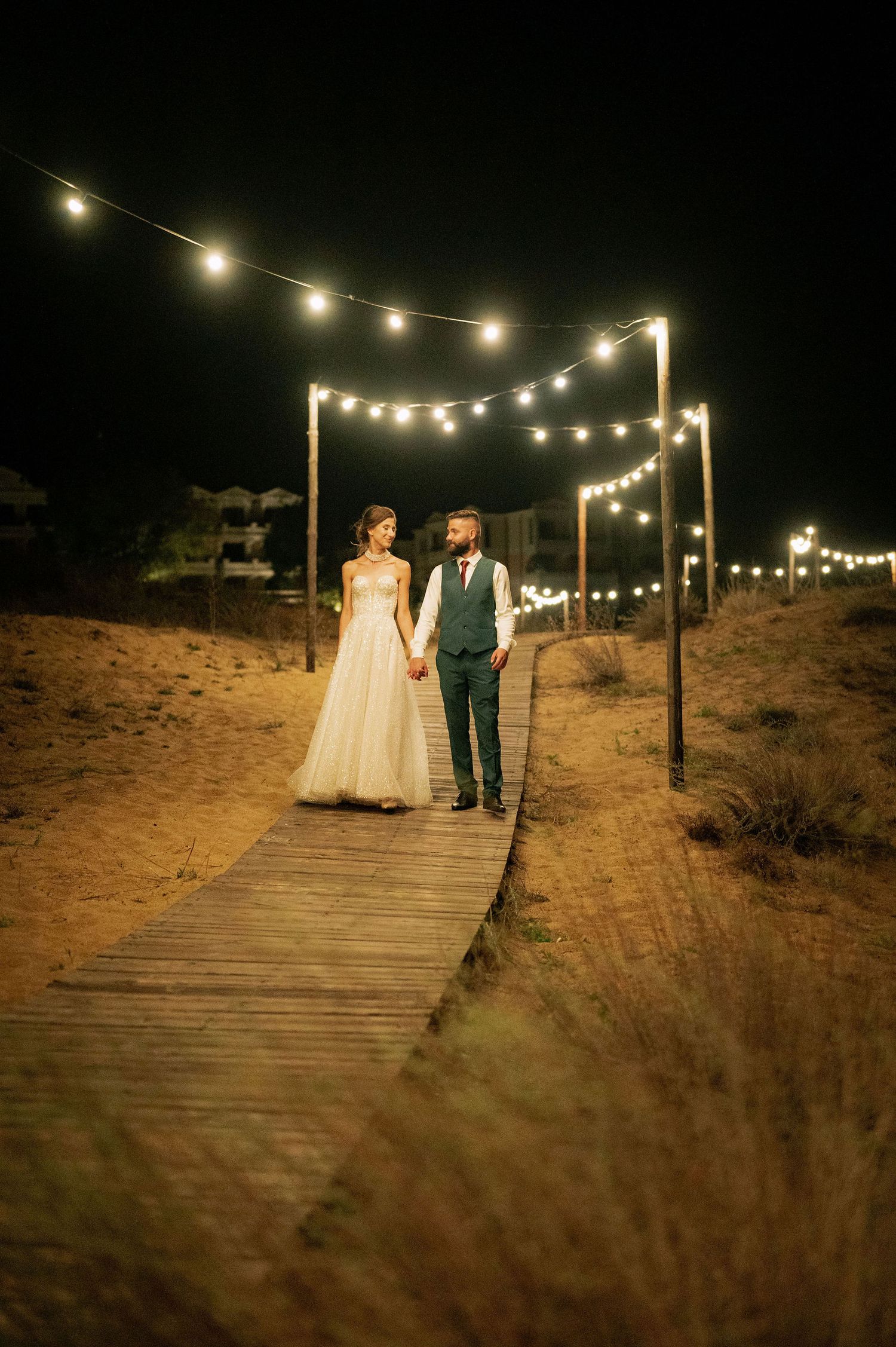 A wedding couple stands hand in hand on a boardwalk illuminated by string lights at dusk.