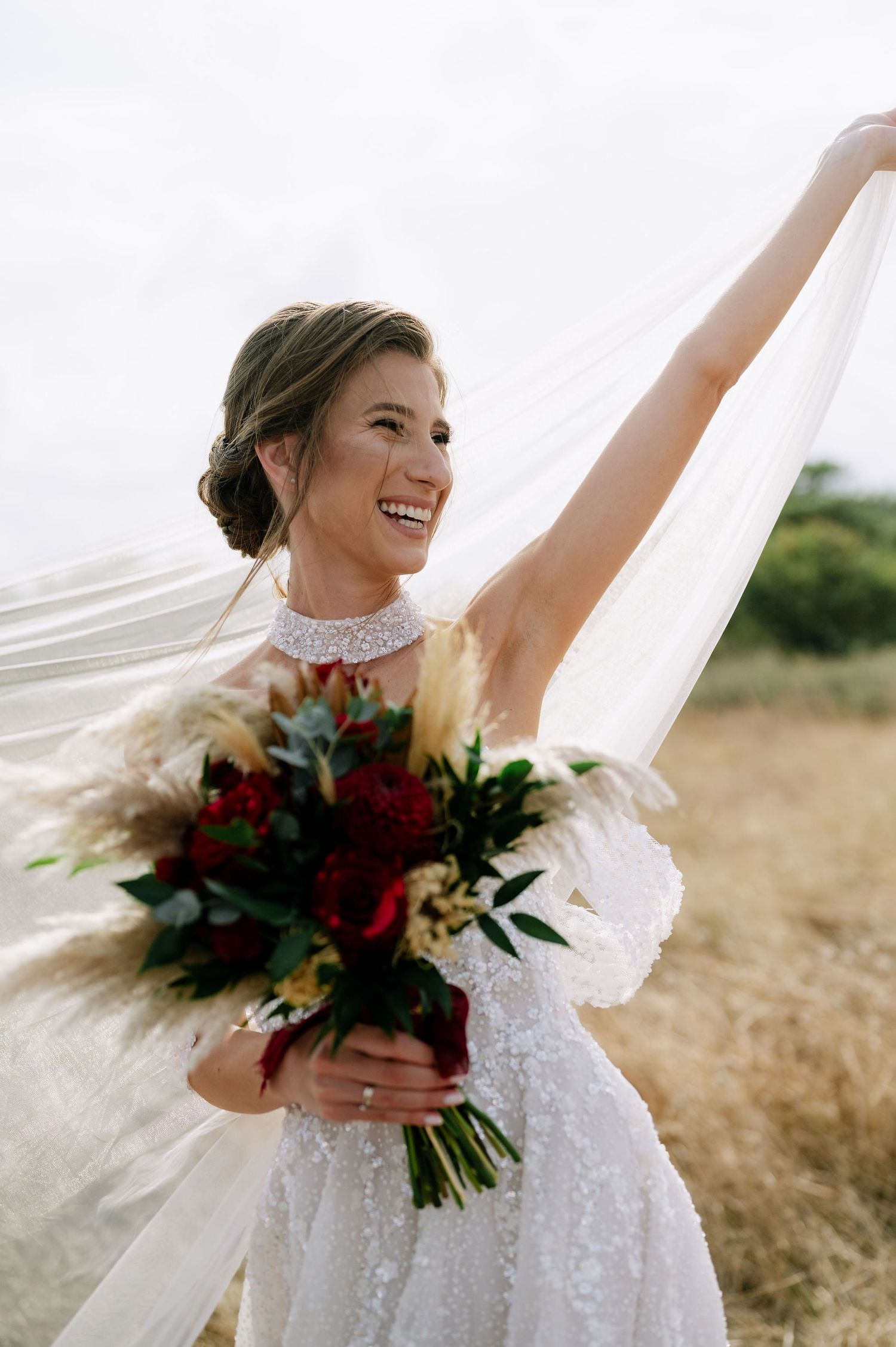 A bride twirls in her white wedding dress while holding a burgundy bouquet against a golden field.
