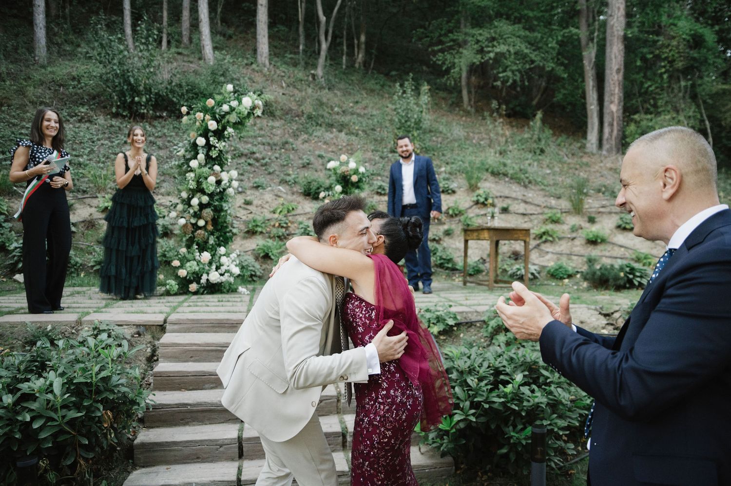 Wedding guests share a joyful moment during an outdoor ceremony with floral arch and stone steps.